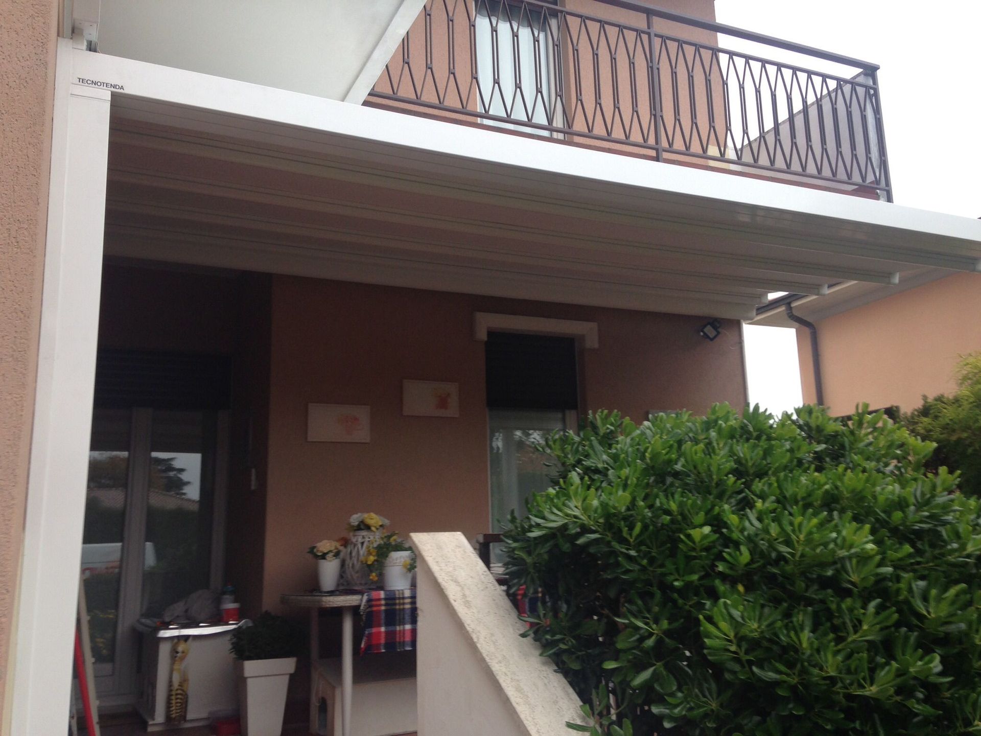 Una tettoia bianca addossata a una casa marrone, accanto a un grande cespuglio e a una scala in pietra.