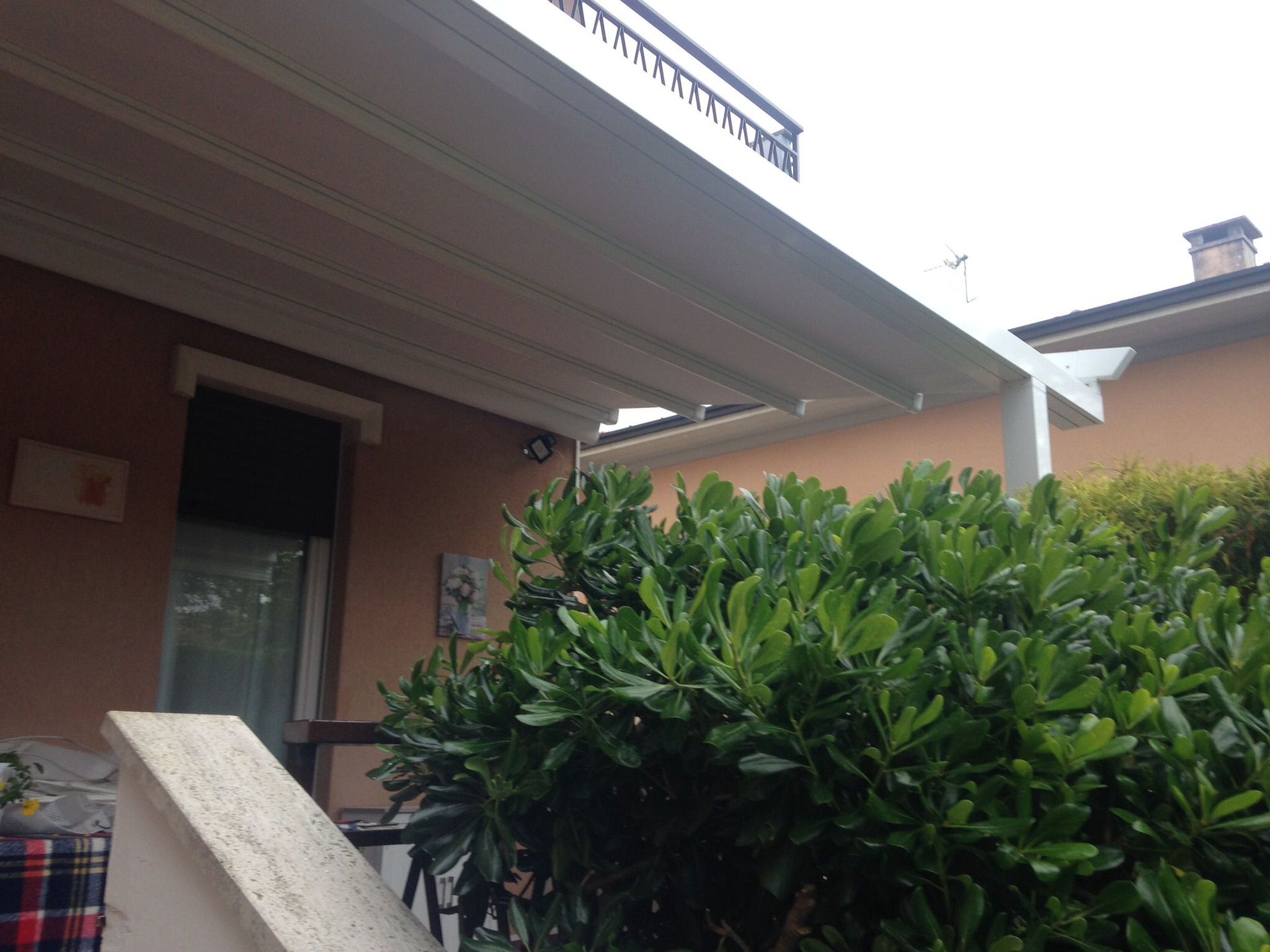 Un patio con una tettoia bianca a pergola sopra un muro della casa e un grande cespuglio verde e frondoso in primo piano.