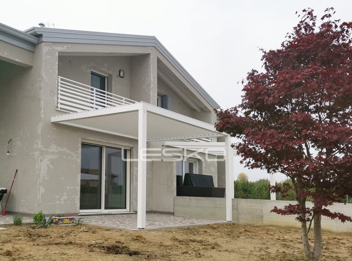 Una casa beige a due piani con un pergolato bianco sopra una porta scorrevole in vetro e un albero dalle foglie rosse in giardino.