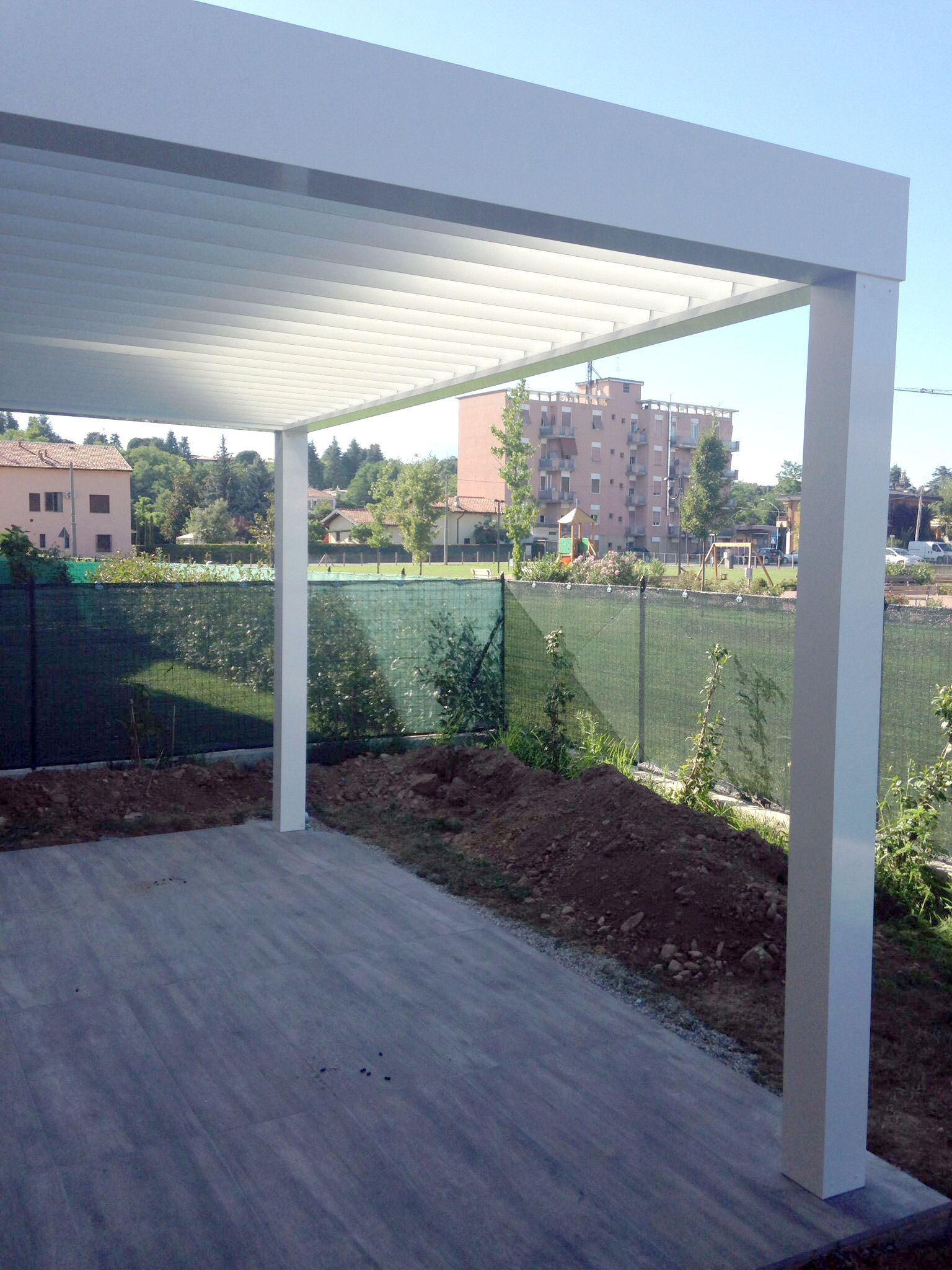Una pergola bianca con lamelle orientabili copre un patio pavimentato, affacciata su un giardino recintato e su un lontano edificio a più piani.