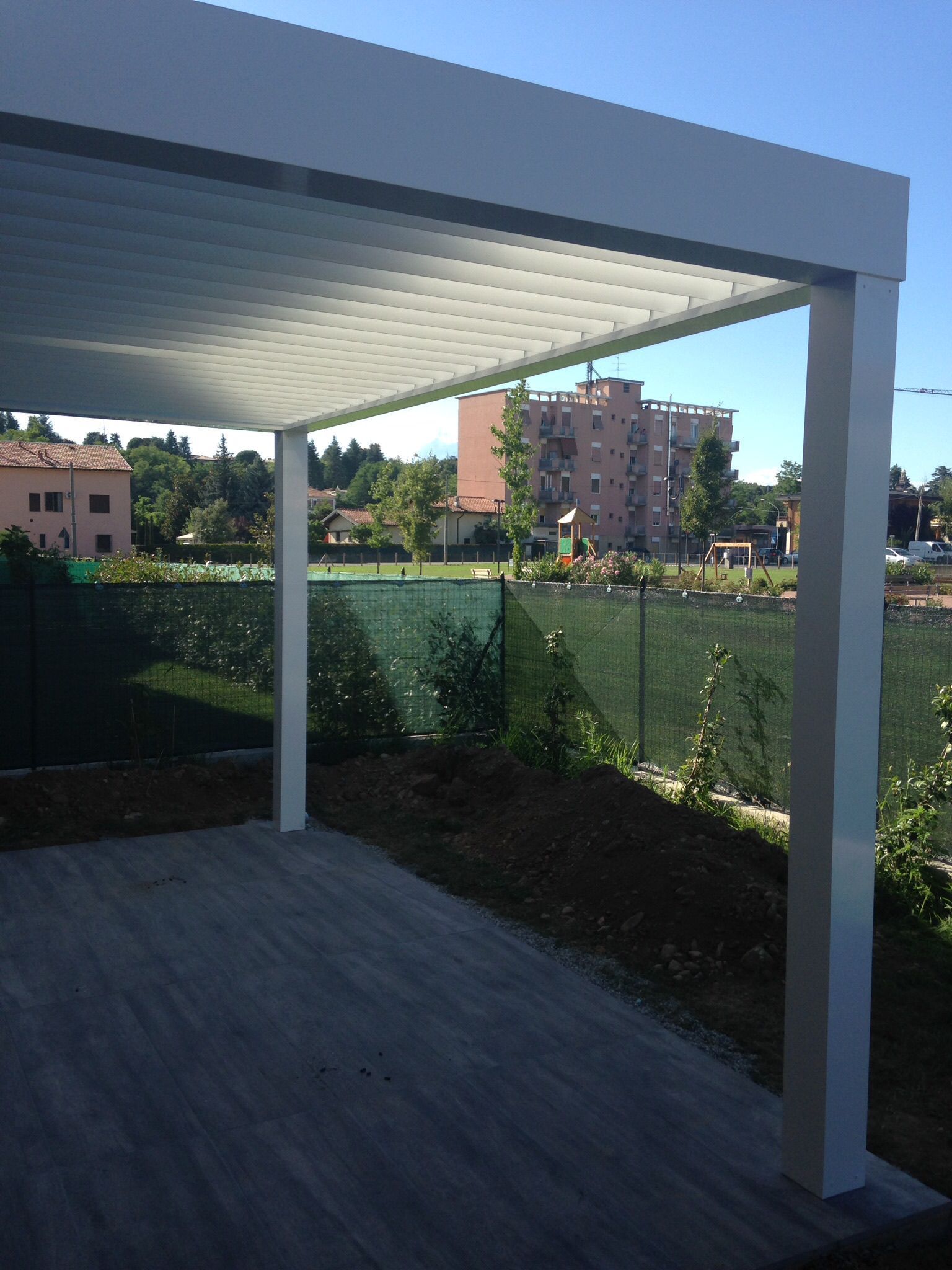 Una moderna pergola bianca con tetto a lamelle regolabili sopra un patio, con vista su un cortile recintato e su un edificio in lontananza.