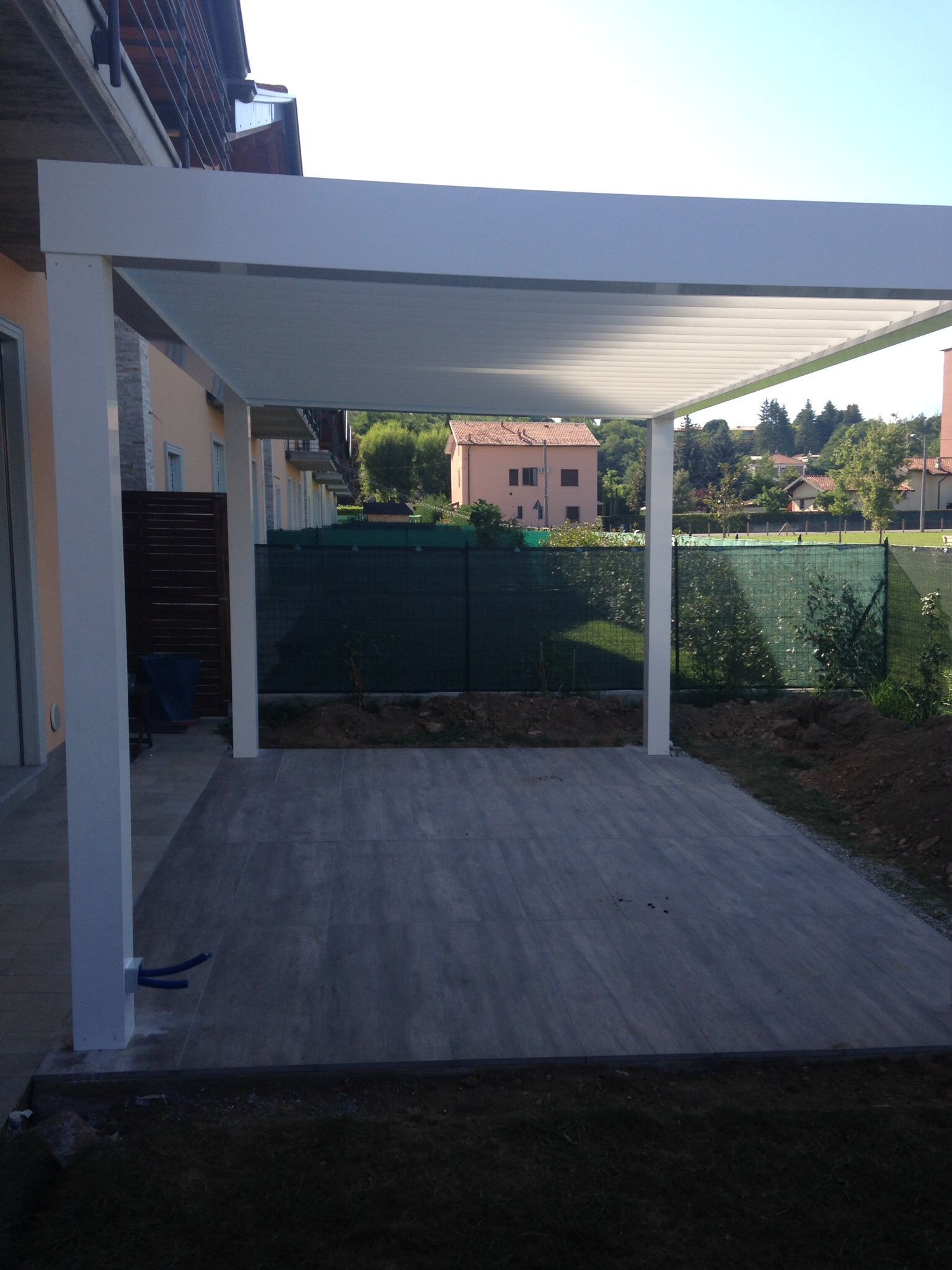 Una moderna pergola bianca con tetto retrattile si erge sopra un patio piastrellato di grigio nel cortile di una casa.