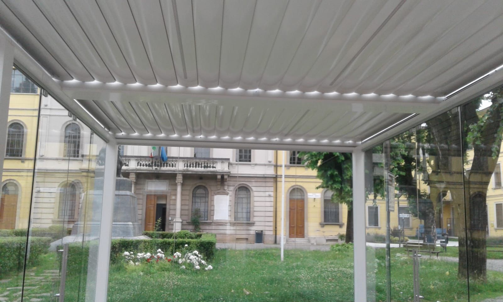 Una struttura di copertura a pergola retrattile di colore bianco si erge in un giardino pubblico di fronte a uno storico edificio giallo.