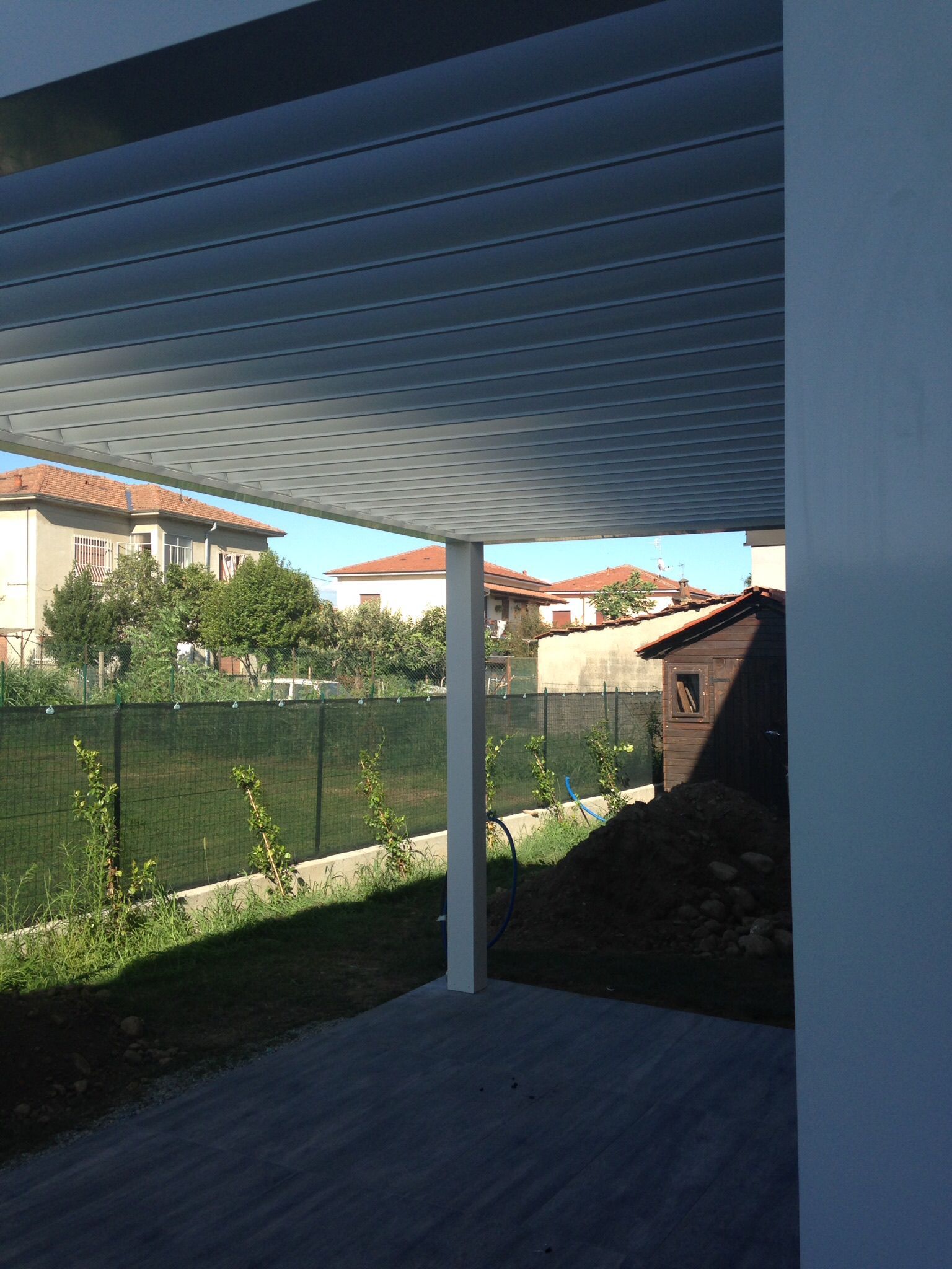 Un pergolato bianco con tetto a doghe sopra un patio pavimentato, affacciato su un giardino con case sullo sfondo.