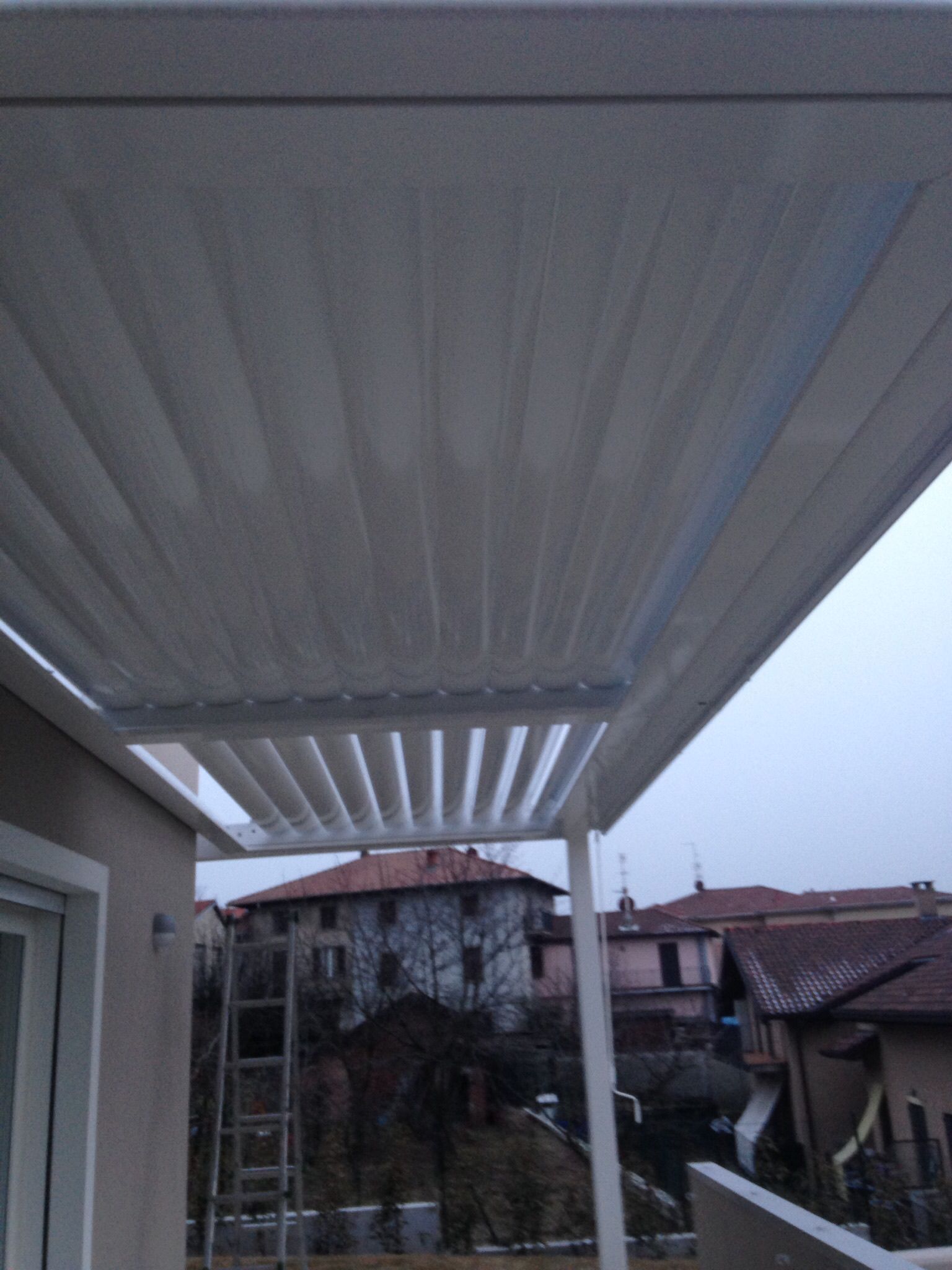 Una pergola a doghe bianche che si estende da una parete di una casa, con vista sugli edifici circostanti sotto un cielo nuvoloso.