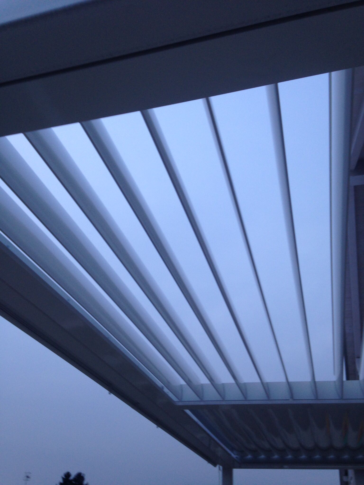 Una vista dal basso di un tetto bianco a lamelle di un patio esterno, con le lamelle parzialmente aperte, sullo sfondo di un cielo blu crepuscolare.