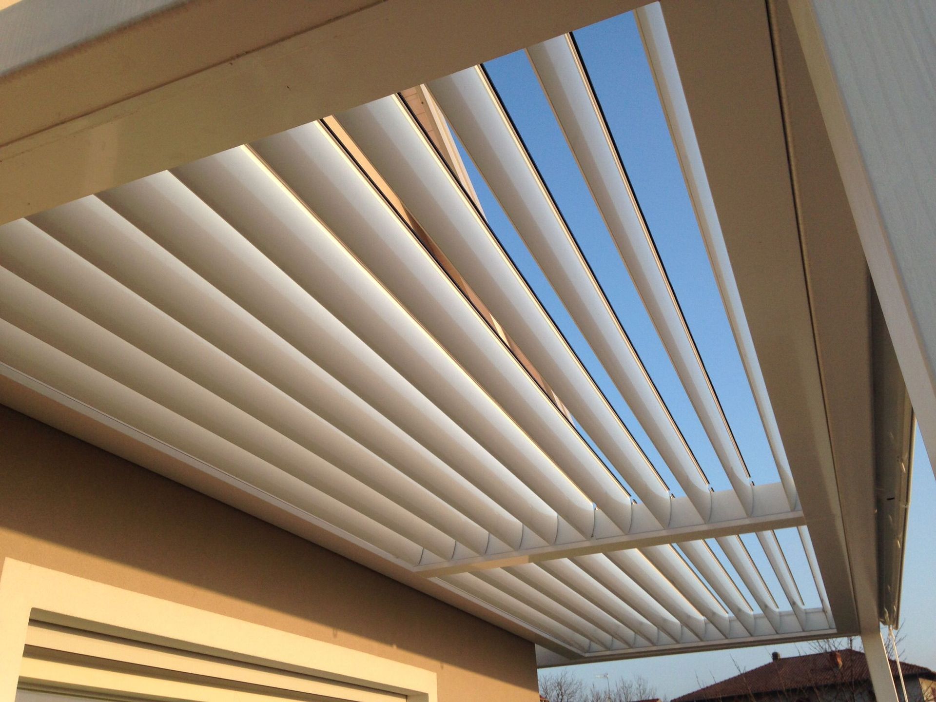 Una tettoia bianca a lamelle regolabili, ideale per il patio, ombreggia l'esterno di un'abitazione sotto un cielo azzurro e limpido.