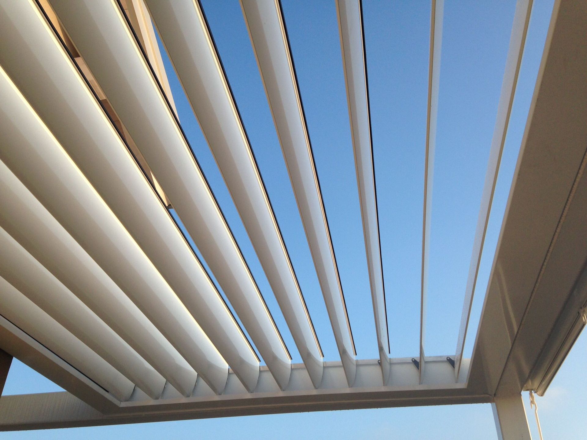 Vista dal basso di un tetto da patio bianco a lamelle regolabili, parzialmente aperto su un cielo azzurro limpido.