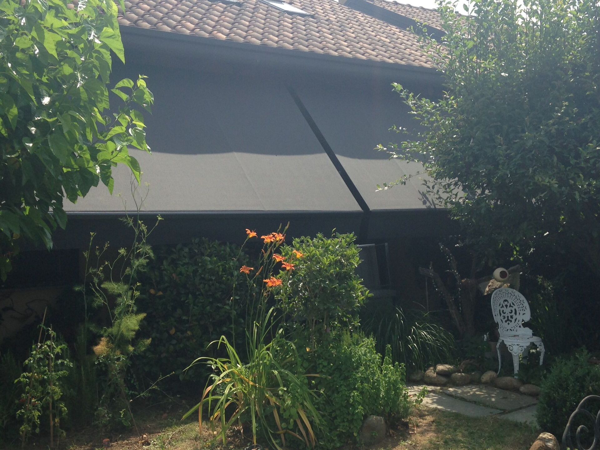 Una tenda da sole scura ombreggia la facciata di un edificio, sul quale si trovano un giardino, dei gigli arancioni e, in primo piano, una sedia ornamentale bianca.