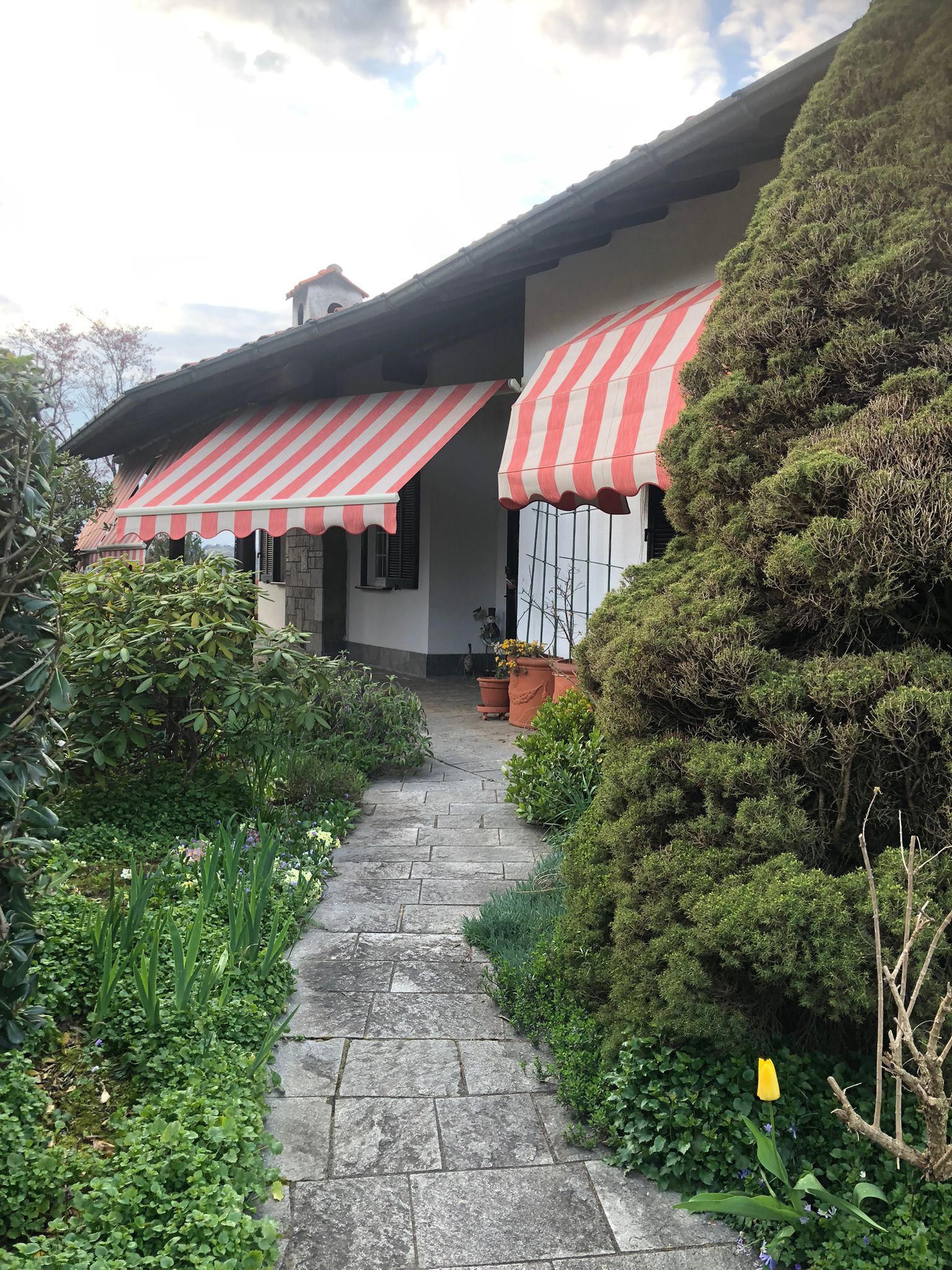 Un sentiero di pietra conduce a una casa con muri bianchi, tende da sole a strisce rosse e bianche e vegetazione circostante.