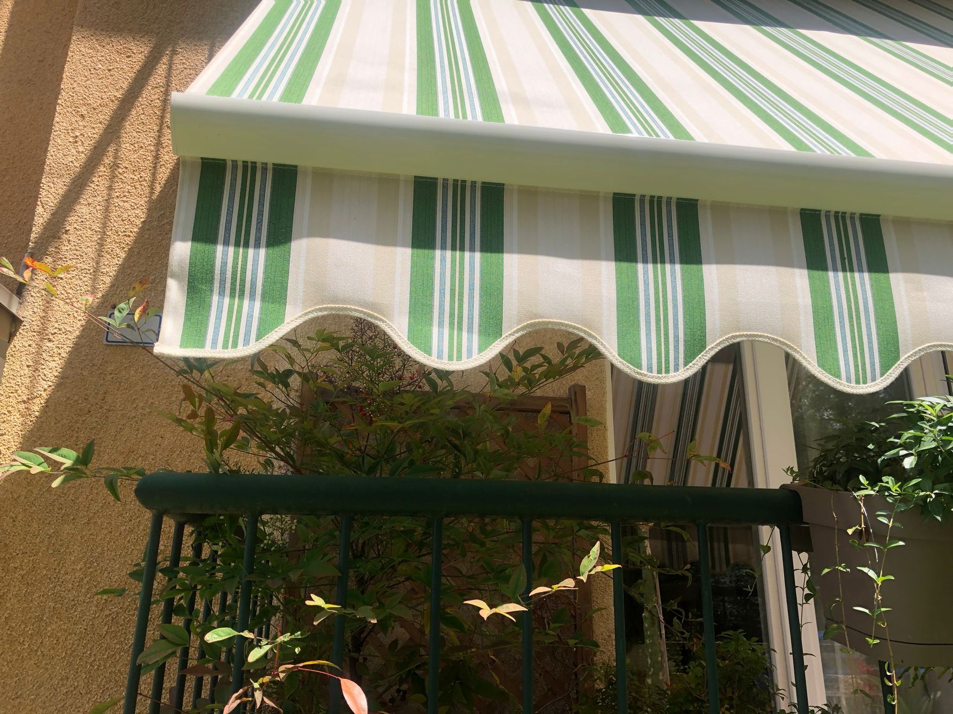 Tenda a righe verdi e beige con bordo smerlato, sovrapposta a una ringhiera e a una vegetazione di colore verde scuro.
