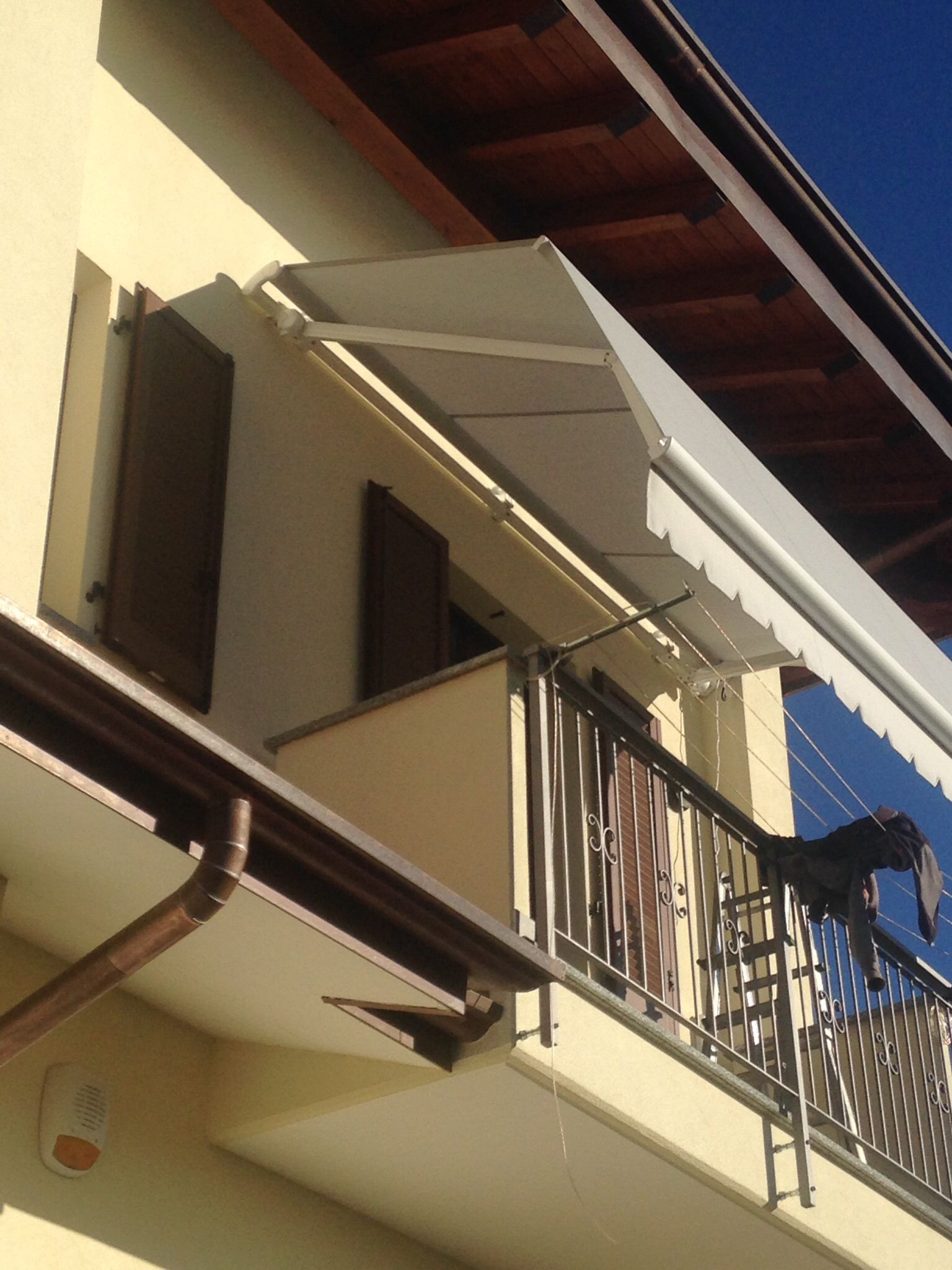 Vista dal basso di un balcone su un edificio di colore chiaro con tenda da sole bianca retrattile e persiane marroni.