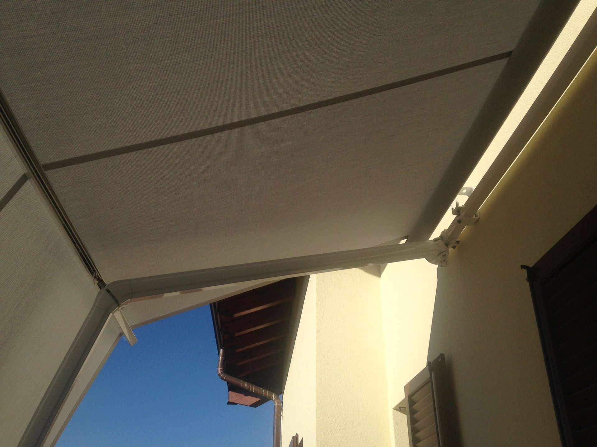 Una tenda da sole in tessuto bianco, fissata a una parete, si estende su un patio con vista parziale sul cielo azzurro e sul tetto della casa.