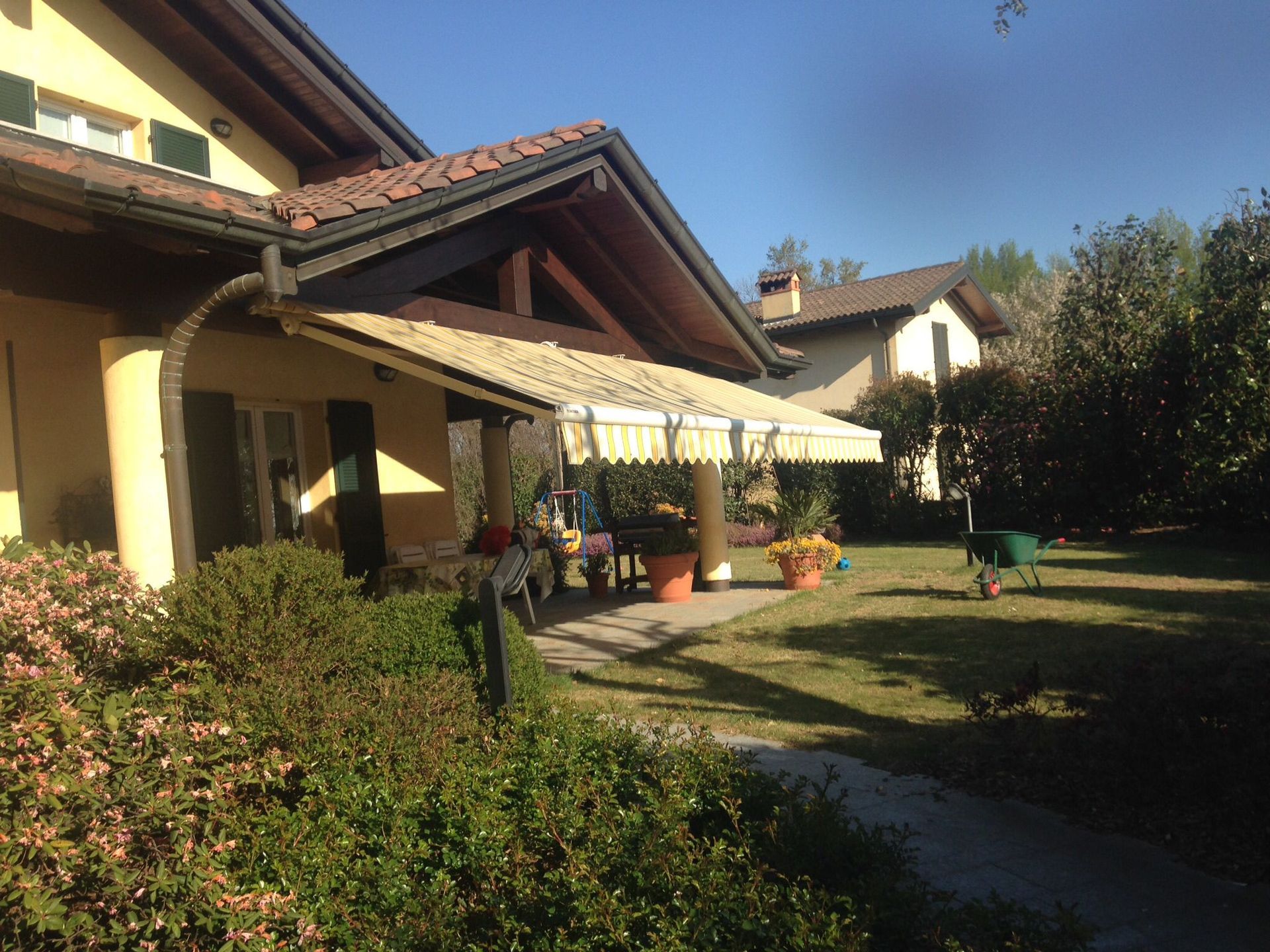 Una casa gialla con un patio coperto, mobili da giardino e un prato sotto un cielo azzurro e limpido.