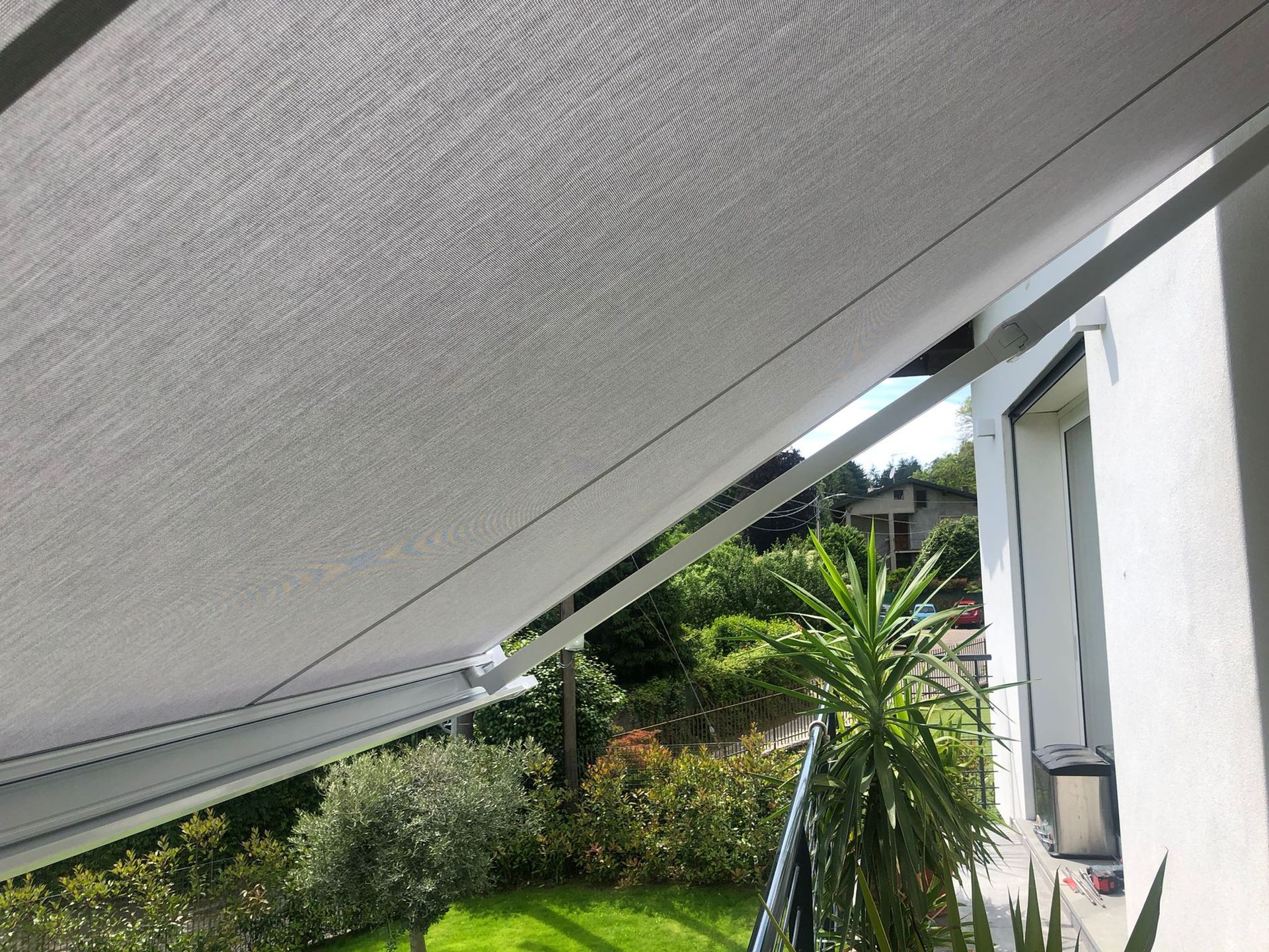 Una vista da sotto una tenda da sole grigio chiaro, che mostra un giardino verde e l'esterno della casa illuminati dalla luce del sole.