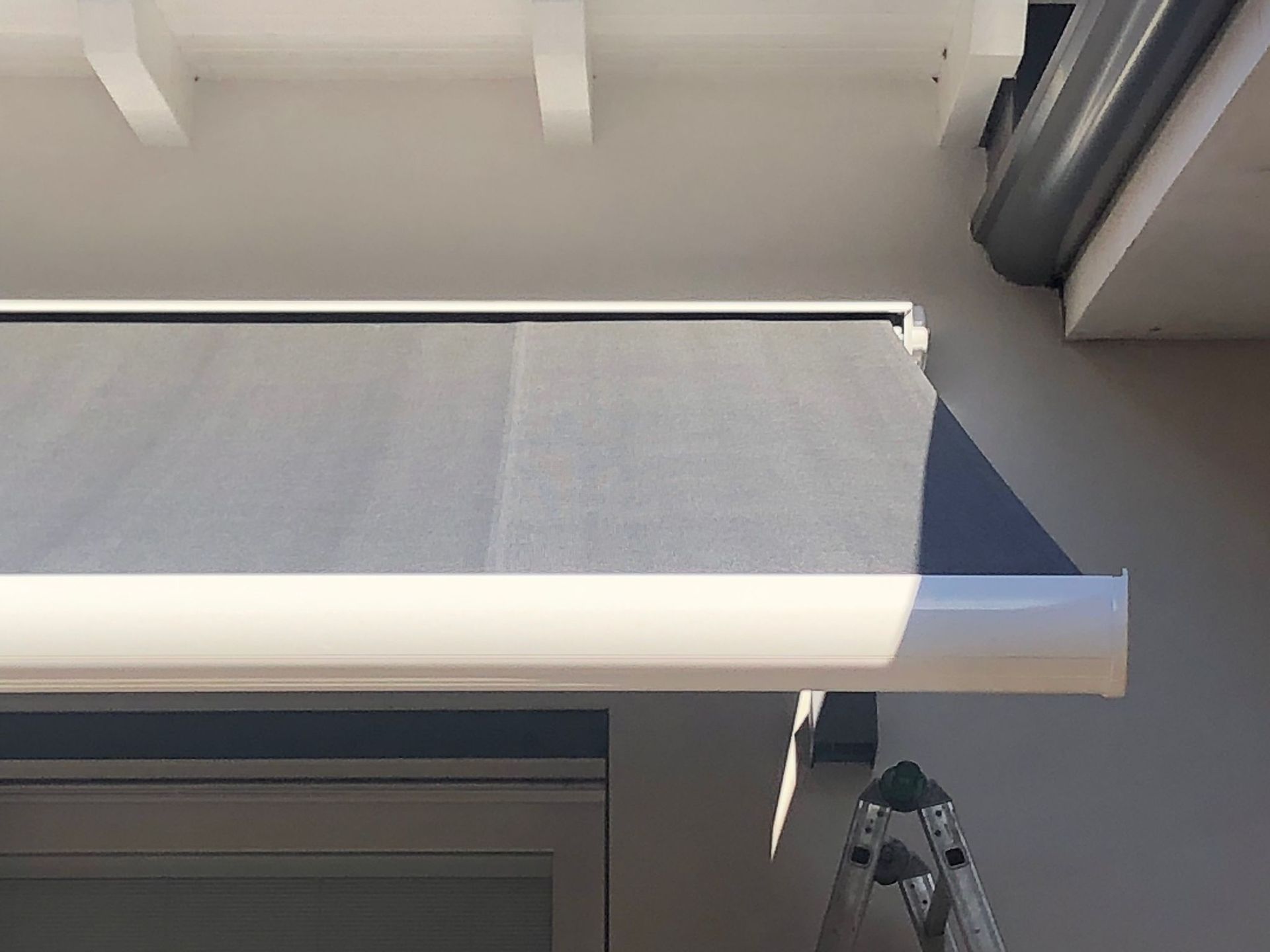 Una tenda da sole retrattile grigia con struttura bianca montata sopra una porta, con la parte superiore di una scala metallica visibile al di sotto.