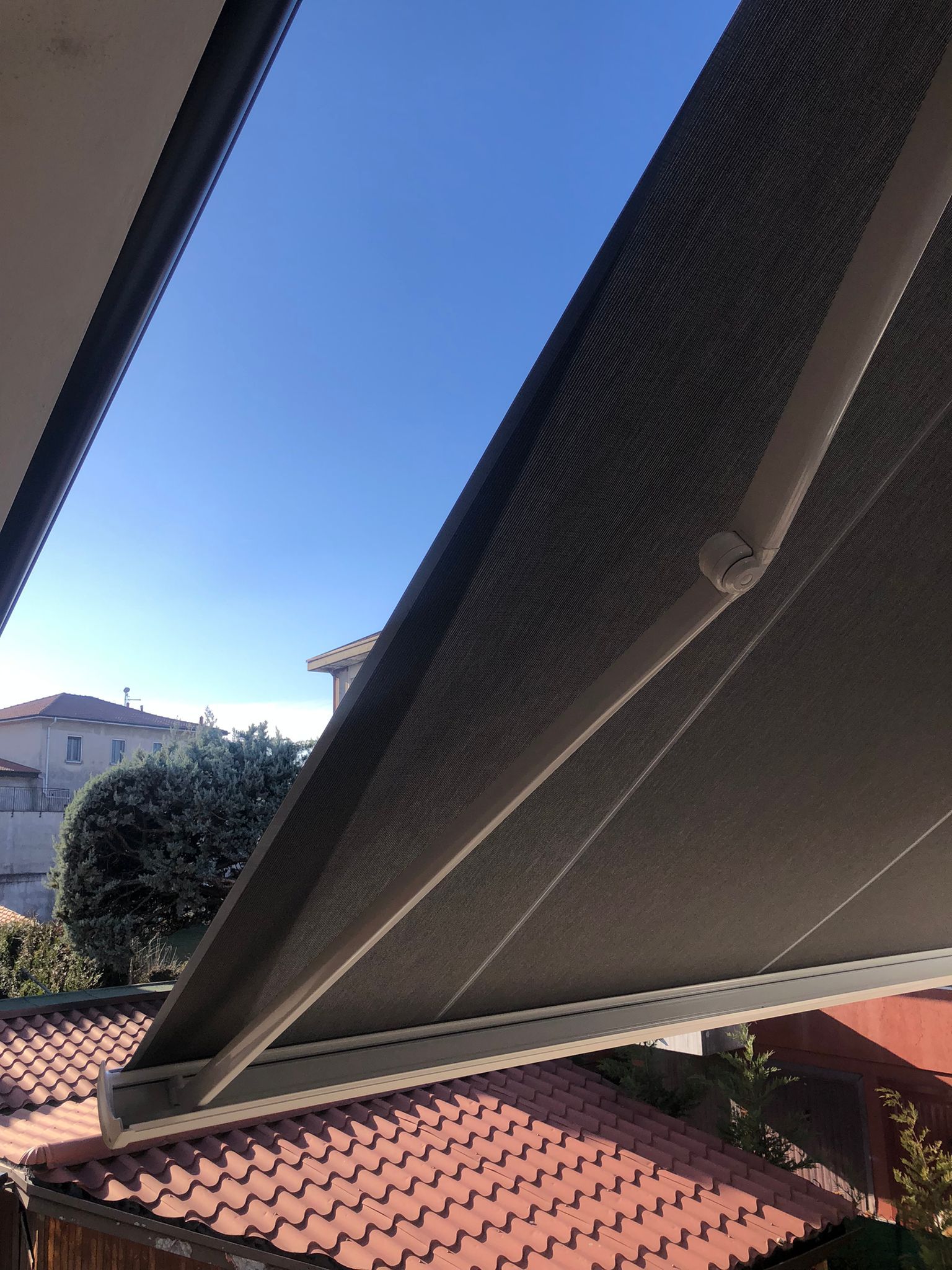 Una vista angolata di una tenda da sole retrattile grigio scuro estesa verso l'esterno, sullo sfondo di un cielo azzurro limpido e di tegole color terracotta.