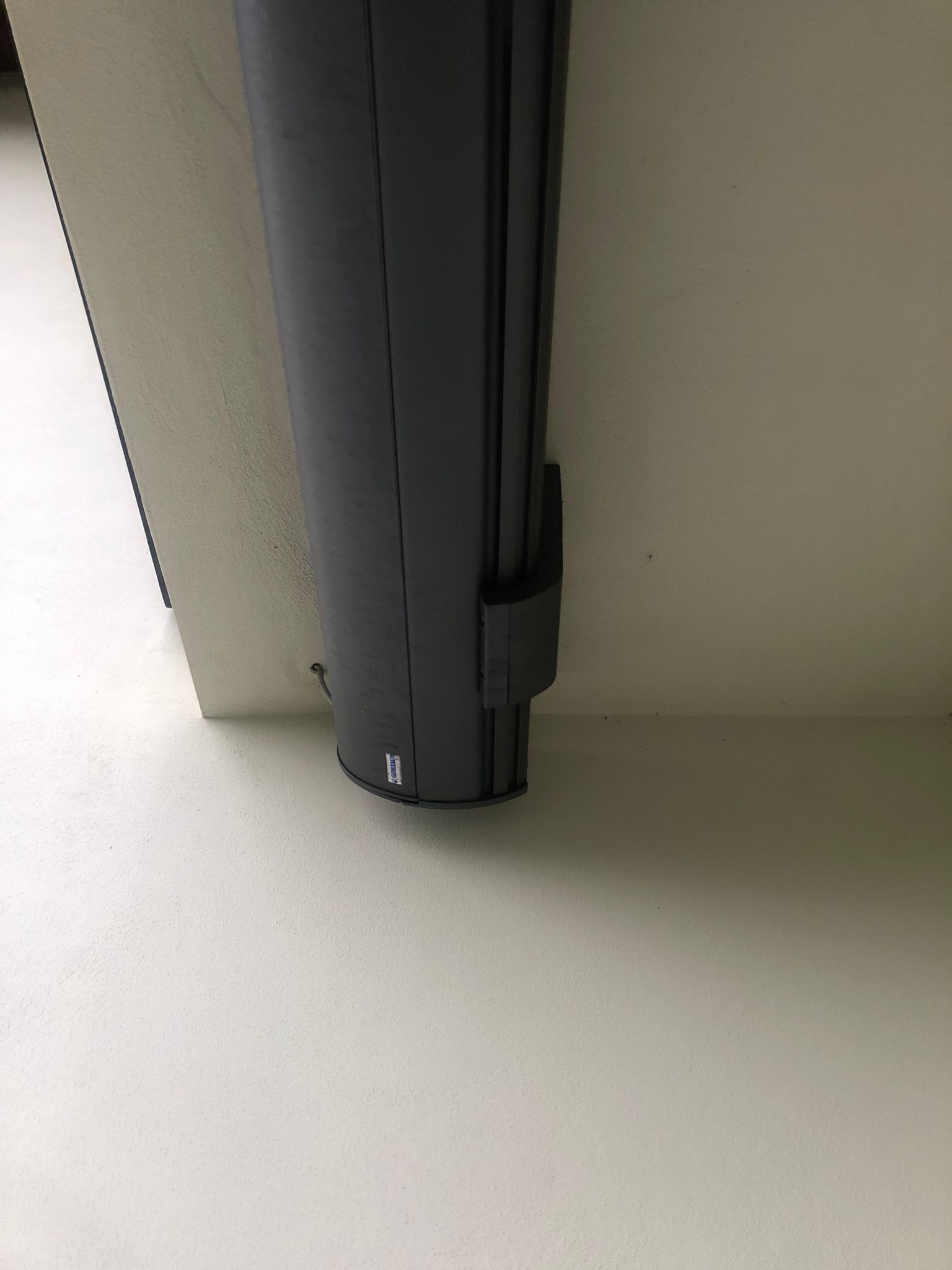 Un meccanismo per tenda a rullo verticale cilindrico di colore nero, fissato a una parete e a un pavimento bianchi.