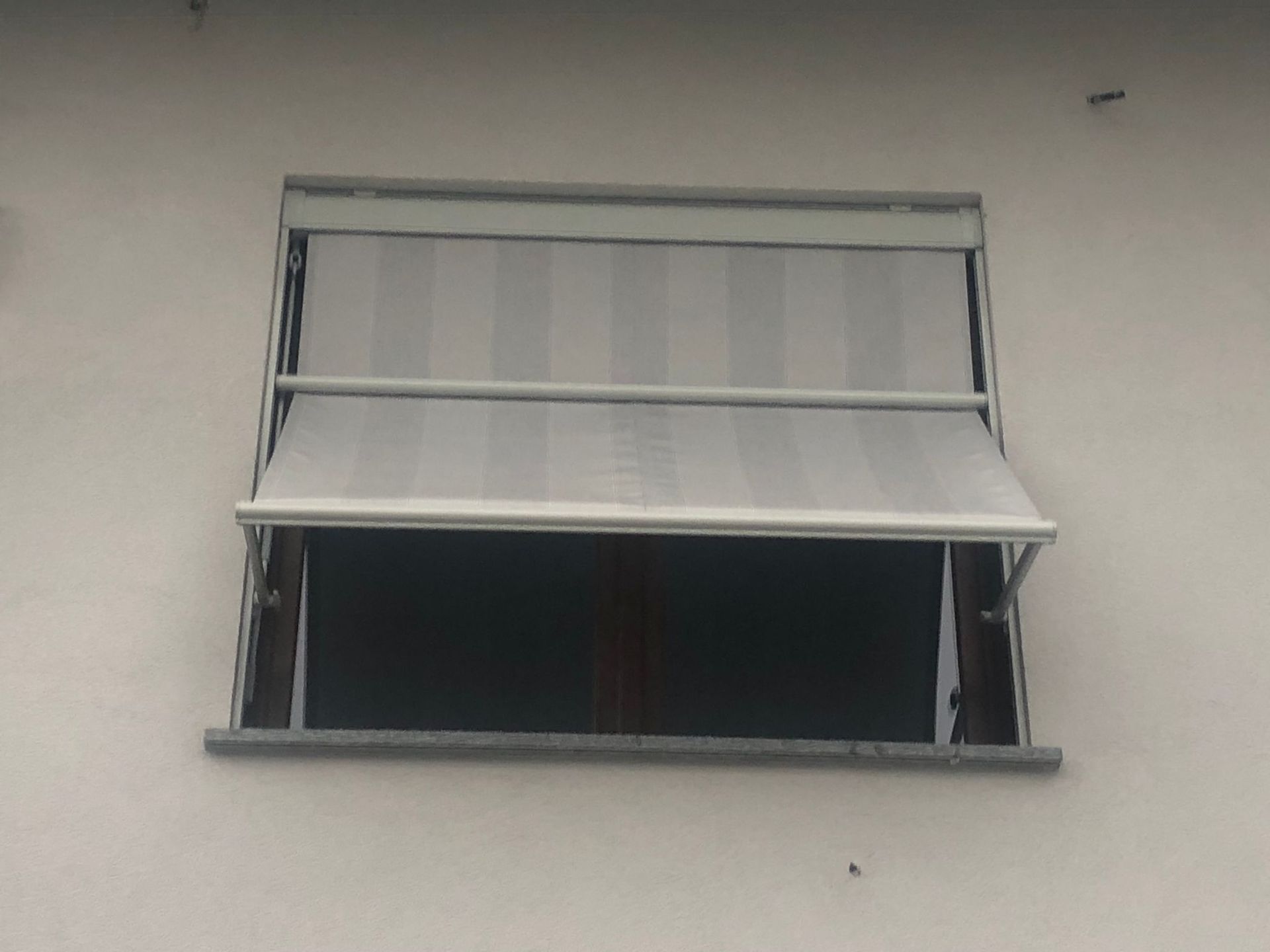 Una finestra con una tenda da sole in tessuto a righe di colore chiaro, parzialmente estesa, stagliata su una semplice parete esterna grigio chiaro.