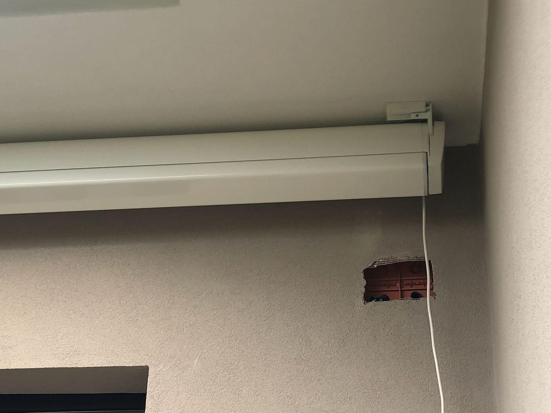 Una tenda bianca montata su un soffitto bianco sopra una parete con una piccola porzione rettangolare di mattoni a vista.