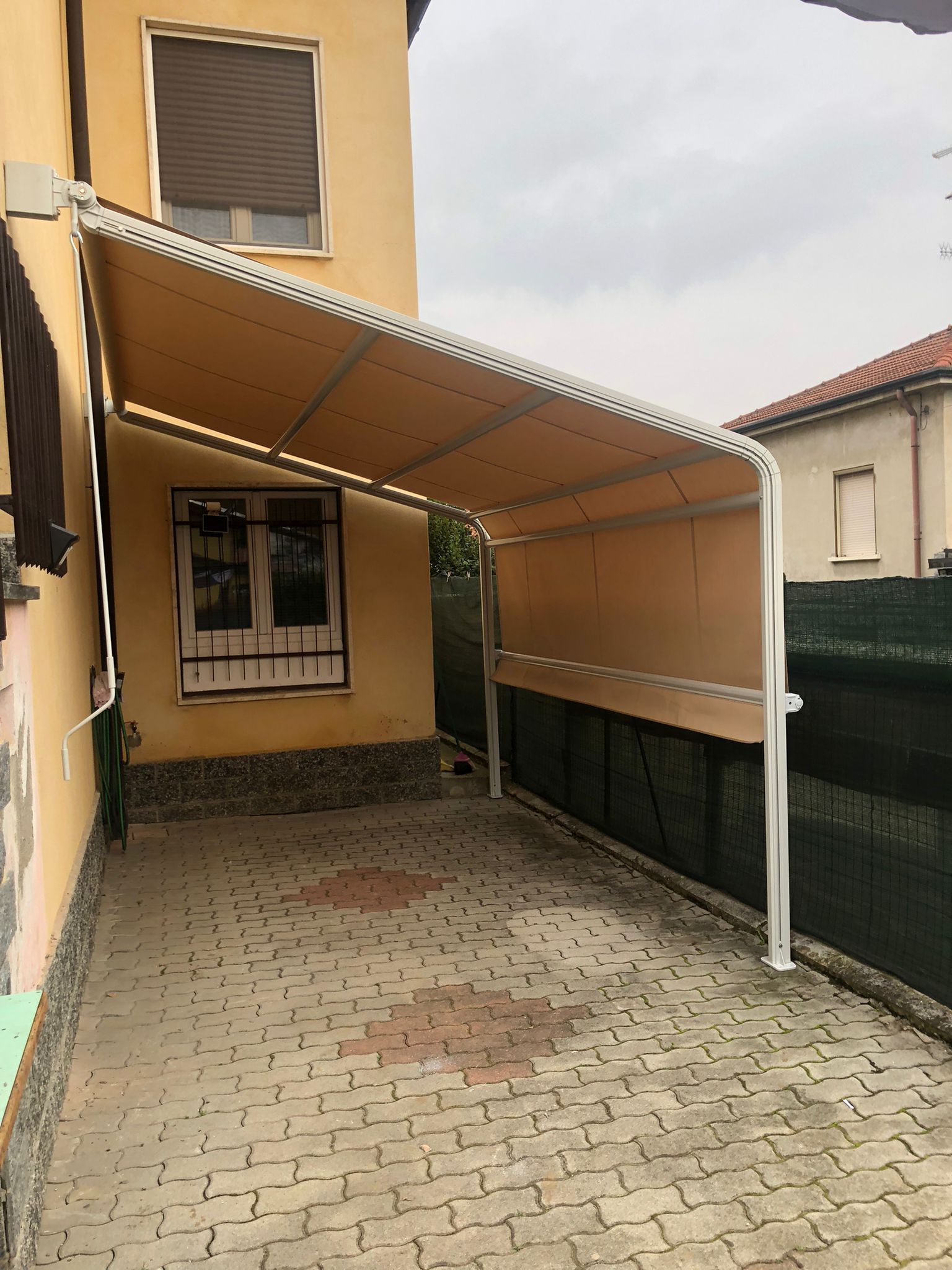 Una tenda da sole retrattile color beige, fissata a una casa gialla, crea uno spazio di parcheggio ombreggiato sopra un patio pavimentato.