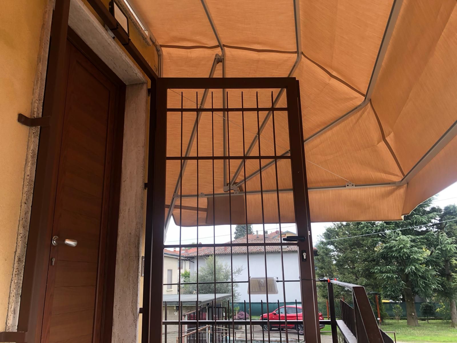 Una porta di legno marrone e un cancello di sicurezza in metallo aperto, visti da sotto una tenda da sole color beige che si affaccia su un giardino.