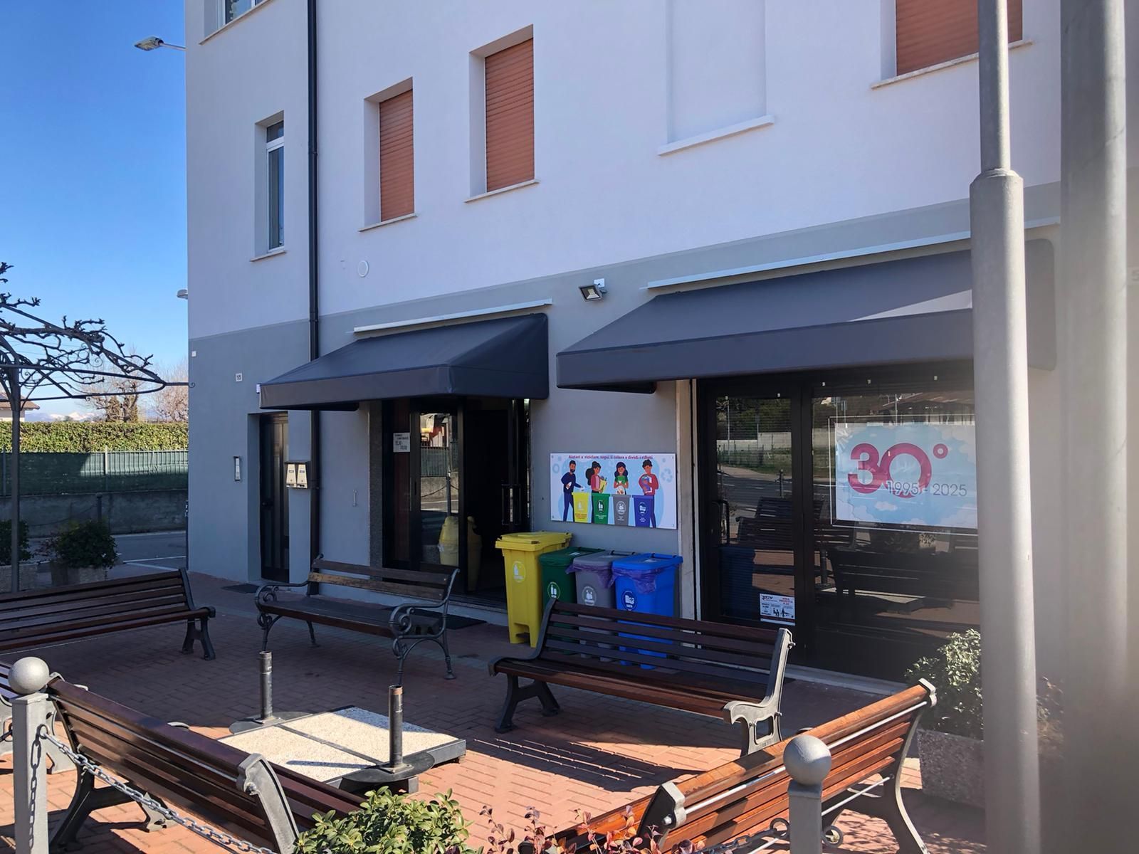 Esterno di un edificio con panchine davanti a due ingressi di negozi e un cartello con la scritta 30° sulla finestra di destra.