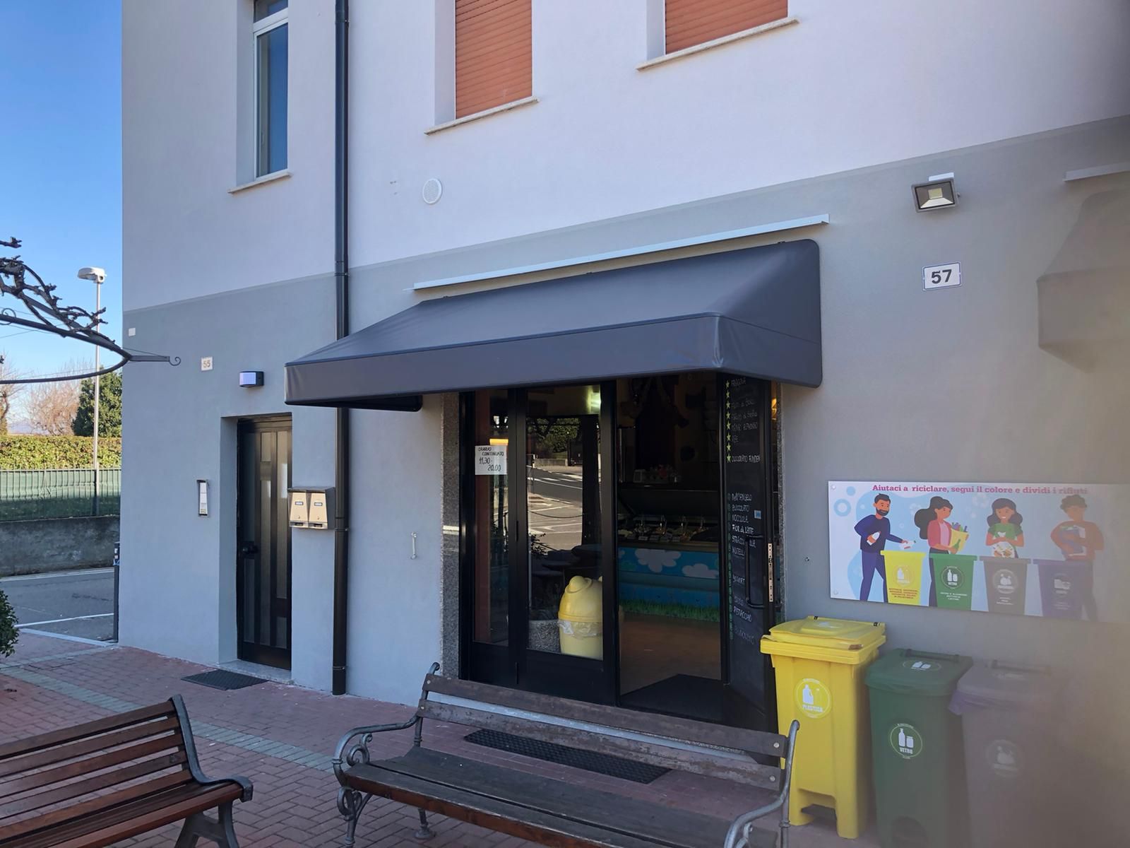 Ingresso di un edificio con una tettoia scura, due panchine e contenitori per la raccolta differenziata, con un cartello a muro che illustra la separazione dei rifiuti.