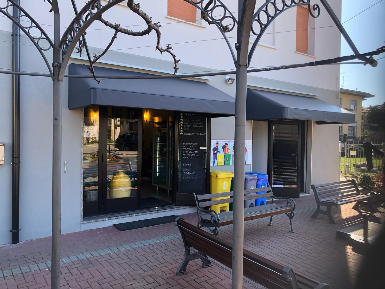 Vetrina con tende da sole grigie, panchine e contenitori per la raccolta differenziata sotto una struttura metallica ad arco su una piazza pavimentata.