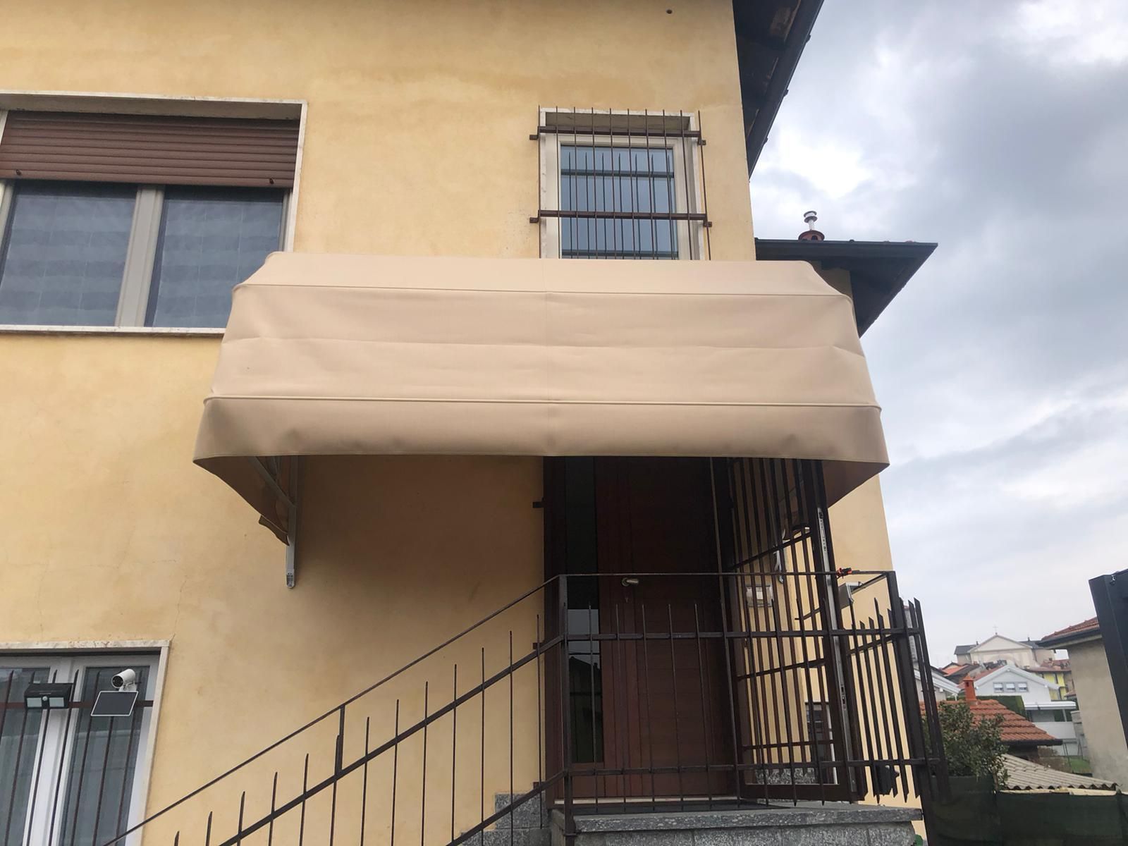 Una tenda da sole in tessuto beige installata sopra la porta d'ingresso di una casa giallo chiaro con inferriate di sicurezza in metallo.