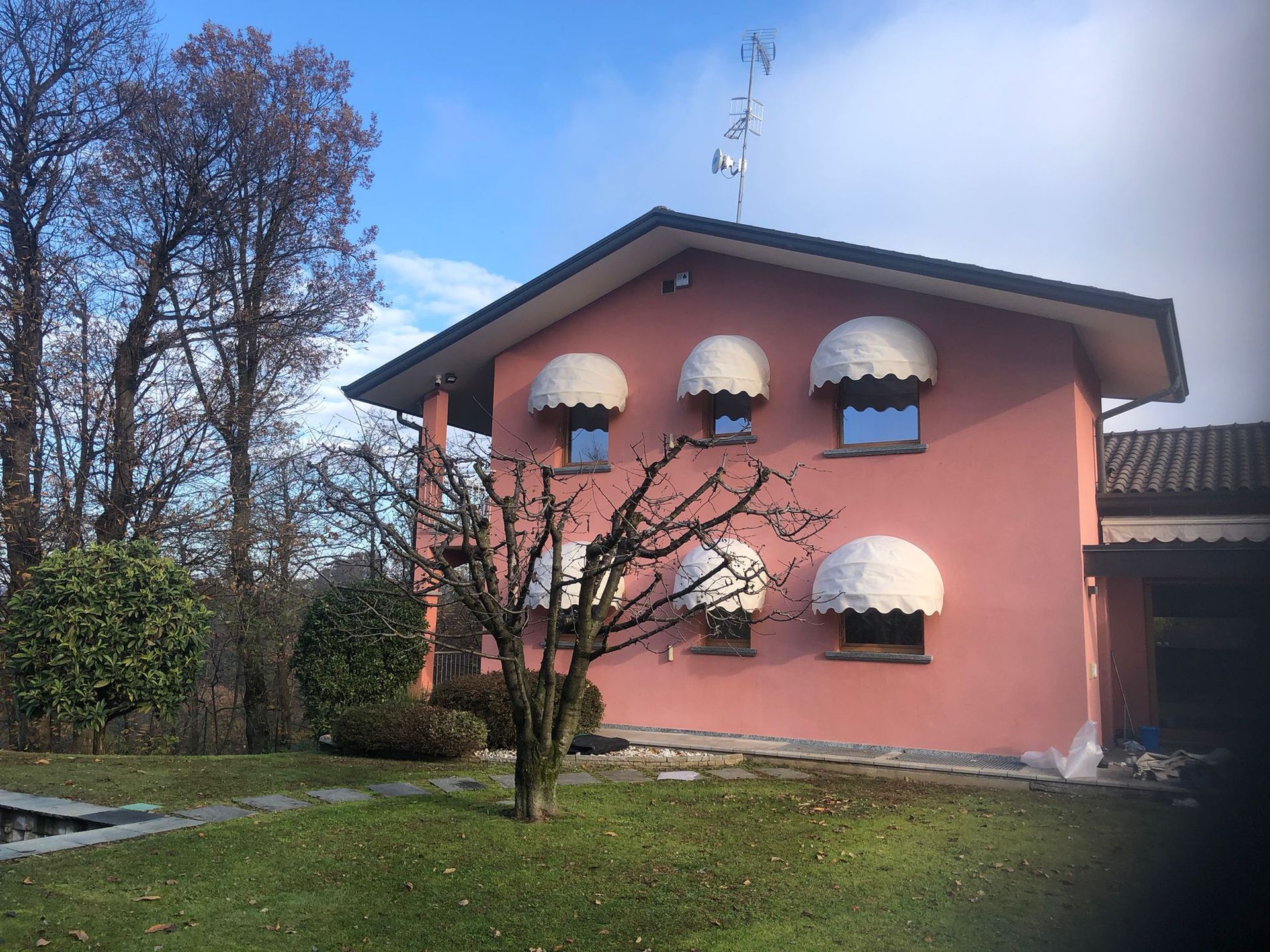 Una casa residenziale rosa a due piani con tetto a due falde, tende da sole bianche alle finestre e un albero spoglio nel giardino anteriore.