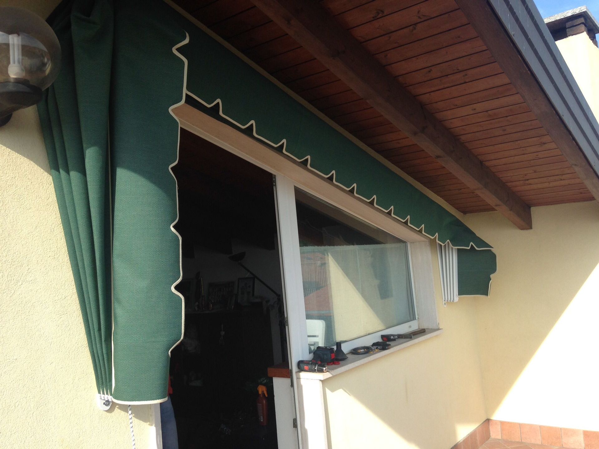 Una tenda da sole retrattile in tessuto verde si estende parzialmente sopra l'ingresso di un patio esterno, al riparo di una tettoia in legno.