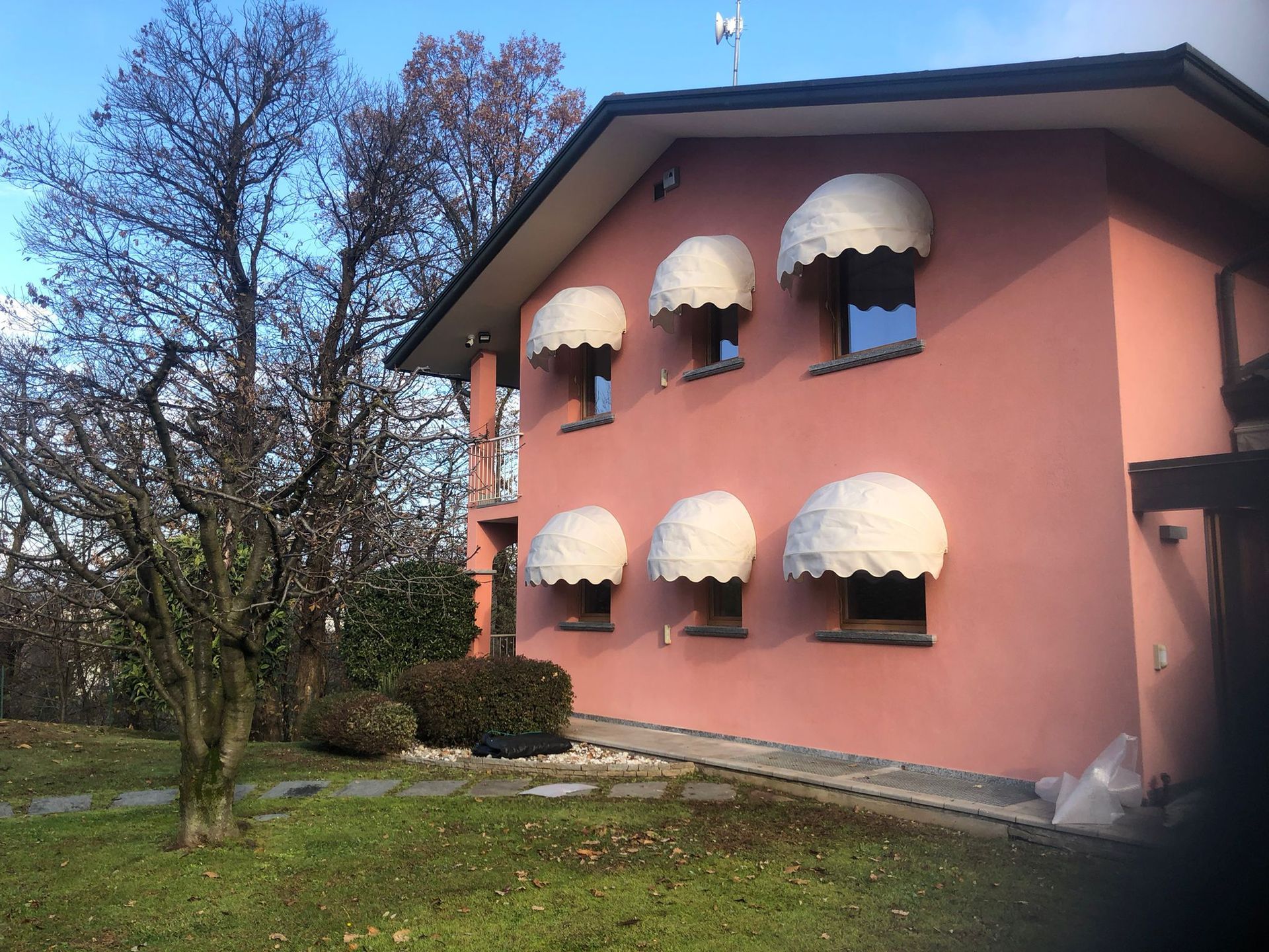 Una casa rosa a due piani presenta sei tende da sole ad arco in tessuto bianco sopra le finestre, circondata da un prato e da alberi.