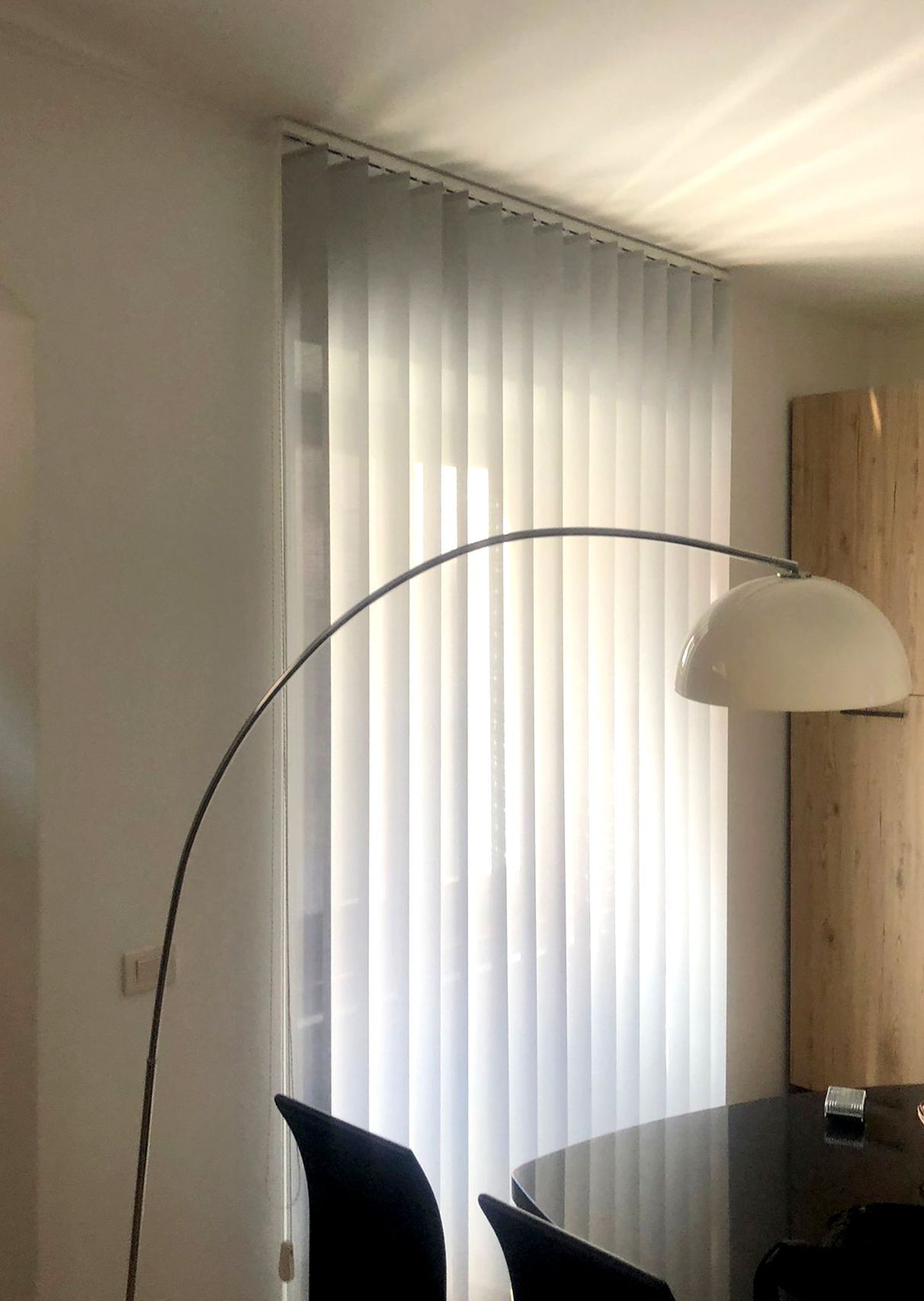 In una stanza con un tavolo scuro, una moderna lampada da terra ad arco è posizionata accanto a una finestra coperta da tende verticali bianche.