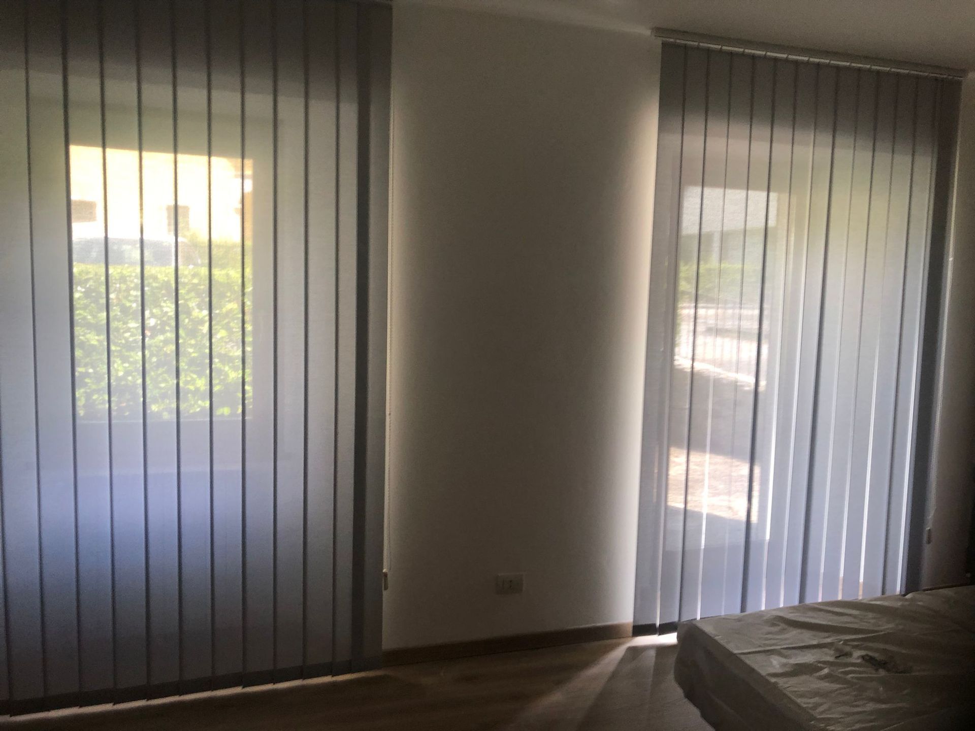 In una stanza minimalista con pavimento in legno, due ampie finestre a tutta altezza coperte da tende verticali in tessuto grigio chiaro.