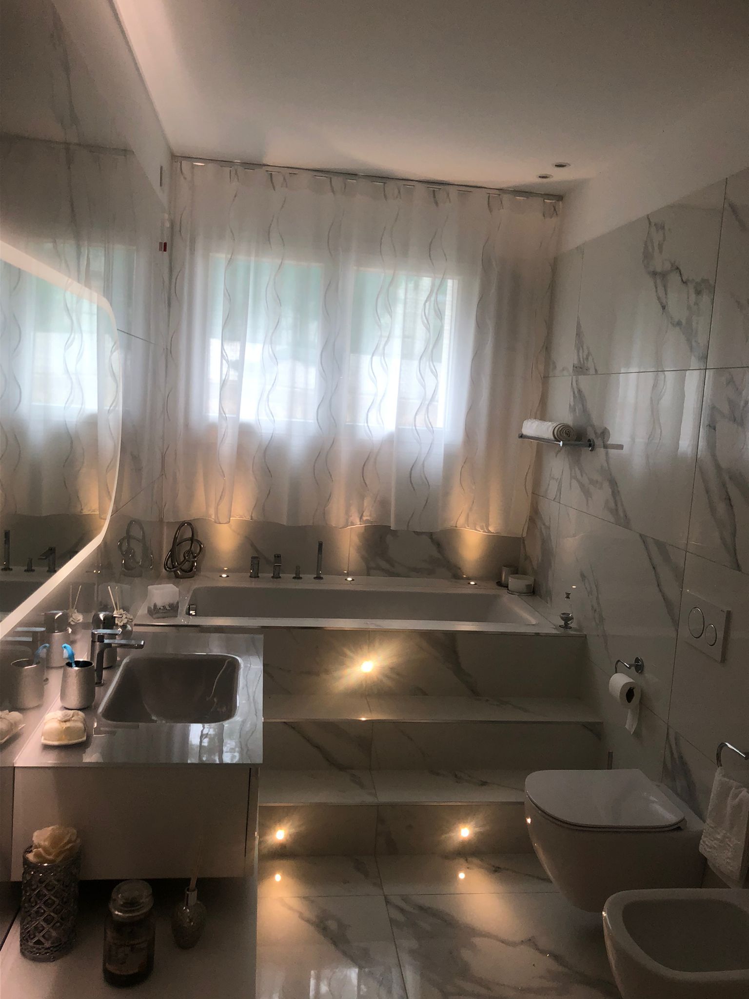 Un bagno in marmo con vasca incassata, lavabo con mobiletto e WC, illuminato da una delicata luce d'accento.