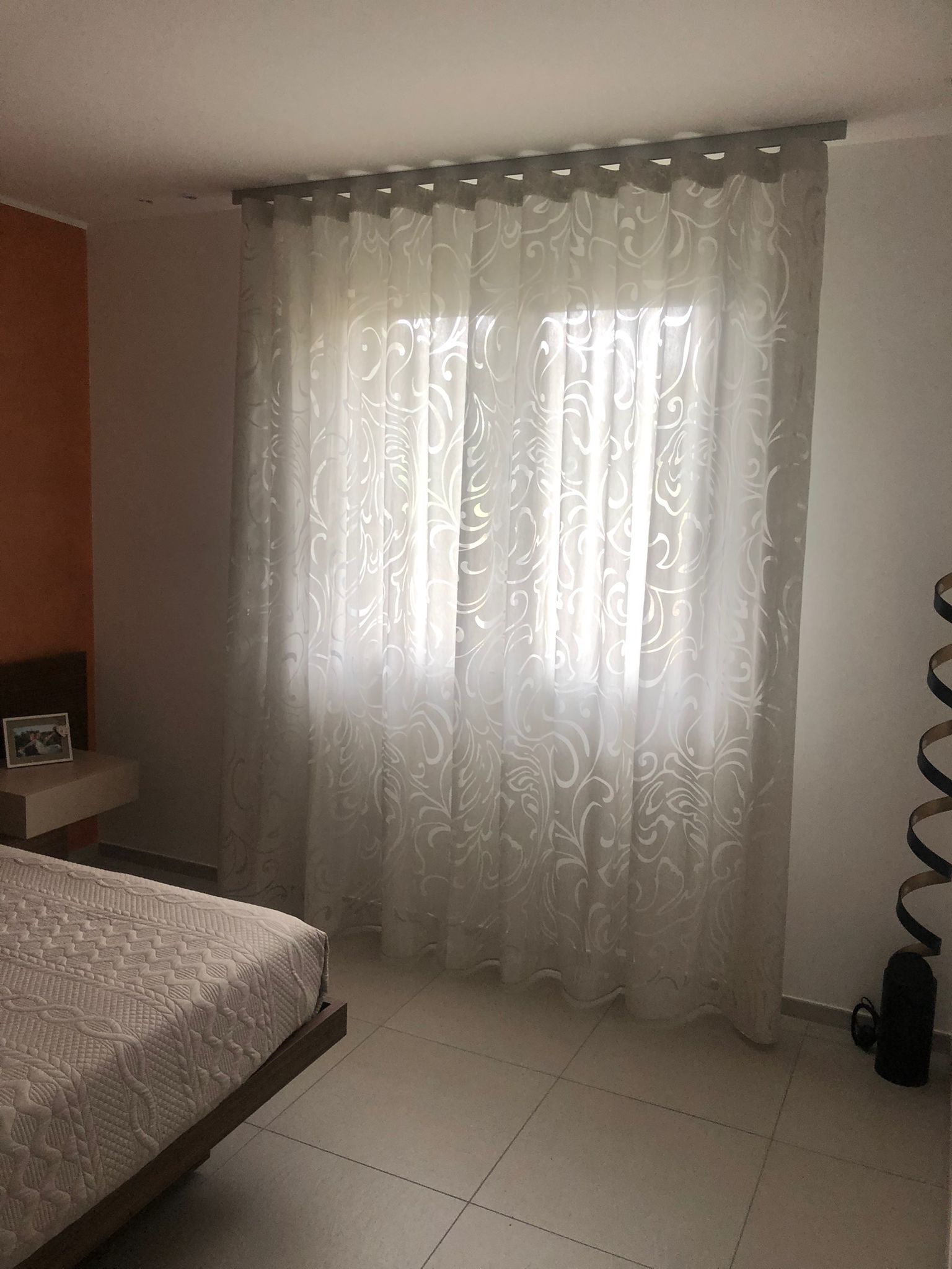 Delle tende di pizzo bianco coprono una finestra di una camera da letto, con un letto visibile in primo piano.