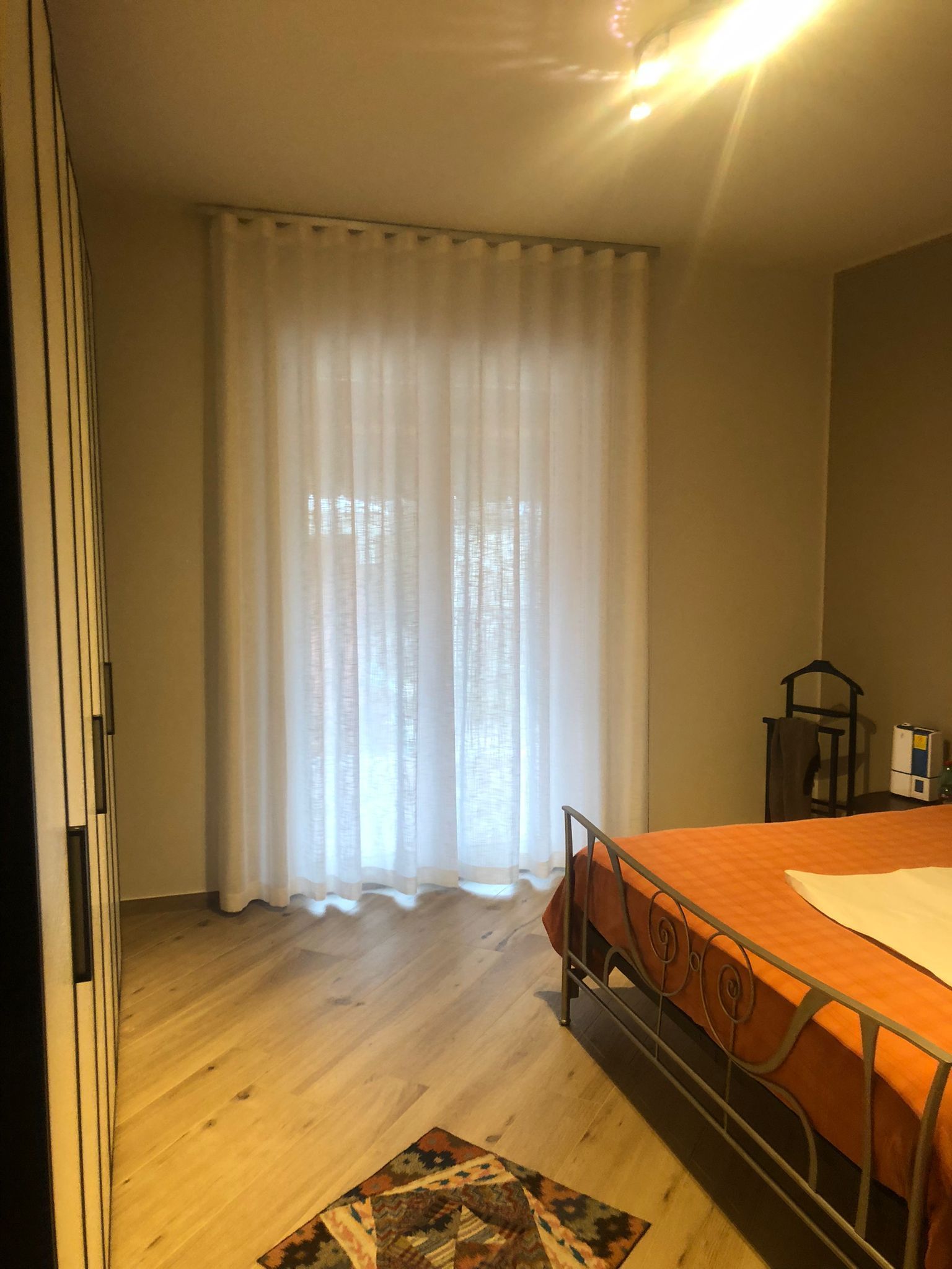 La camera da letto presenta pavimenti in legno chiaro, tende bianche trasparenti sopra la porta-finestra del balcone e un letto con una coperta arancione.