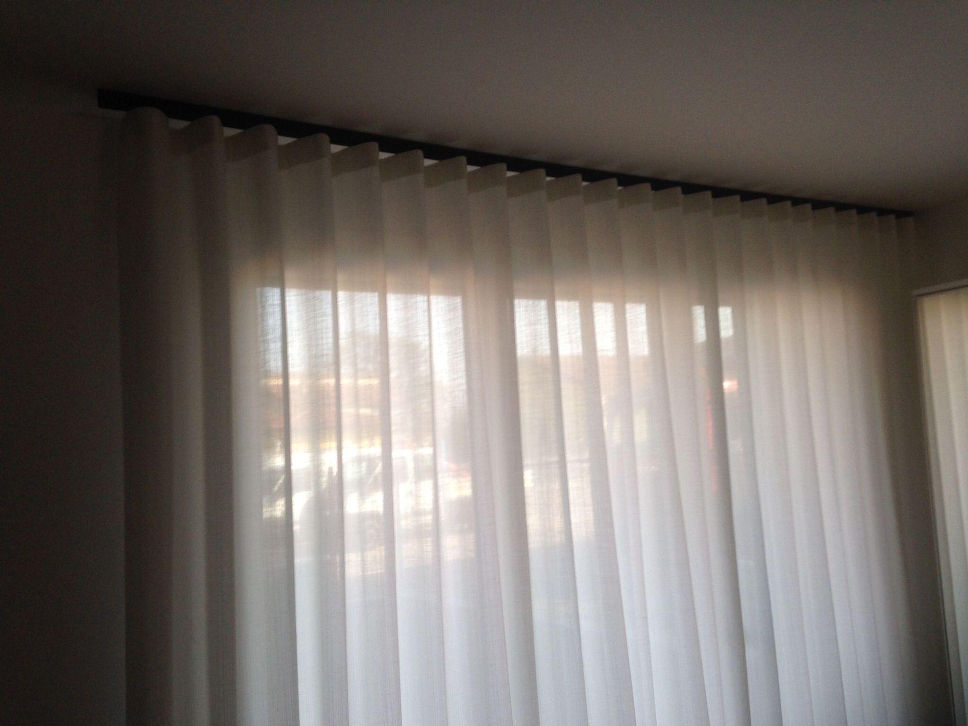 Delle tende bianche trasparenti pendono da un bastone scuro davanti a una finestra, creando morbide onde verticali.