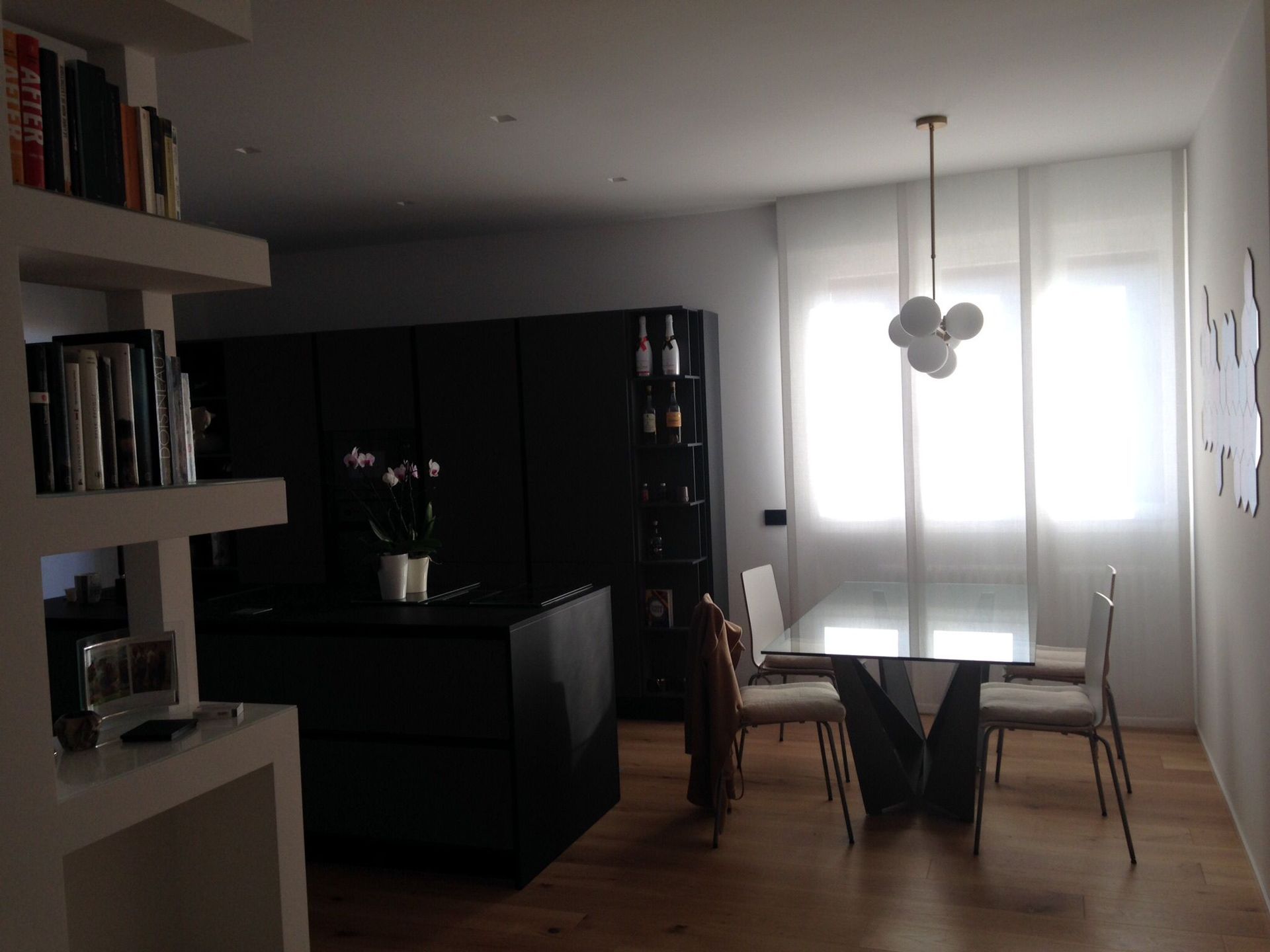 In un luminoso interno domestico, la zona pranzo è arredata con un moderno tavolo in vetro, quattro sedie, una lampada a sospensione a forma di sfera e mobili scuri.