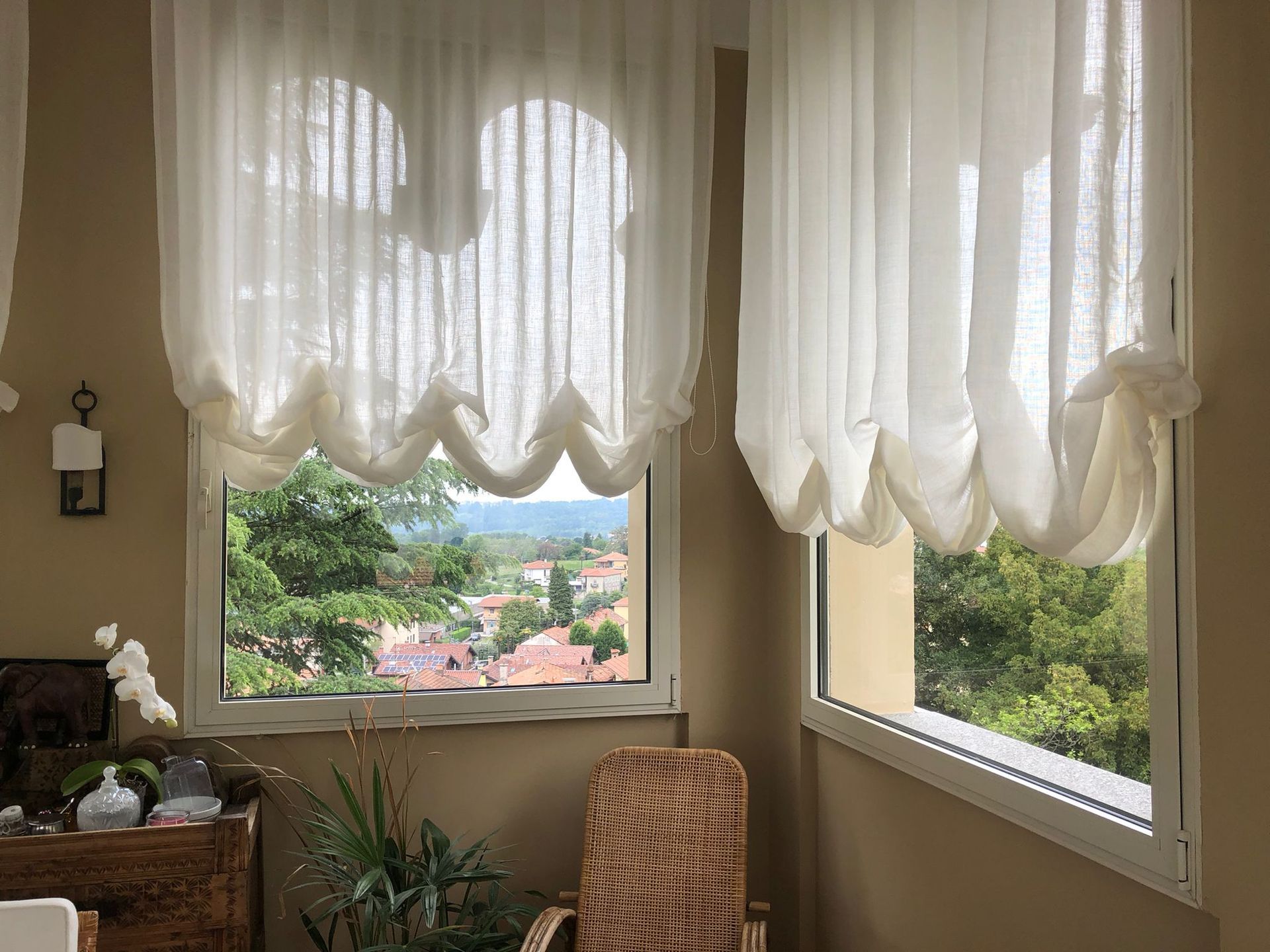 Delle tende bianche e trasparenti coprono due finestre che si affacciano su un paesaggio con una sedia di vimini e una pianta sottostanti.