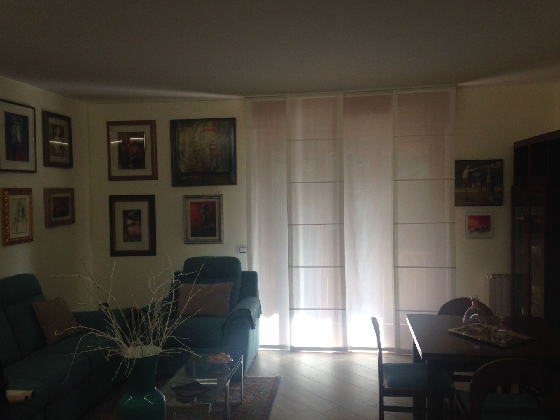 Un accogliente soggiorno con quadri incorniciati alle pareti, un divano blu scuro e una zona pranzo, illuminata da tende luminose alle finestre.