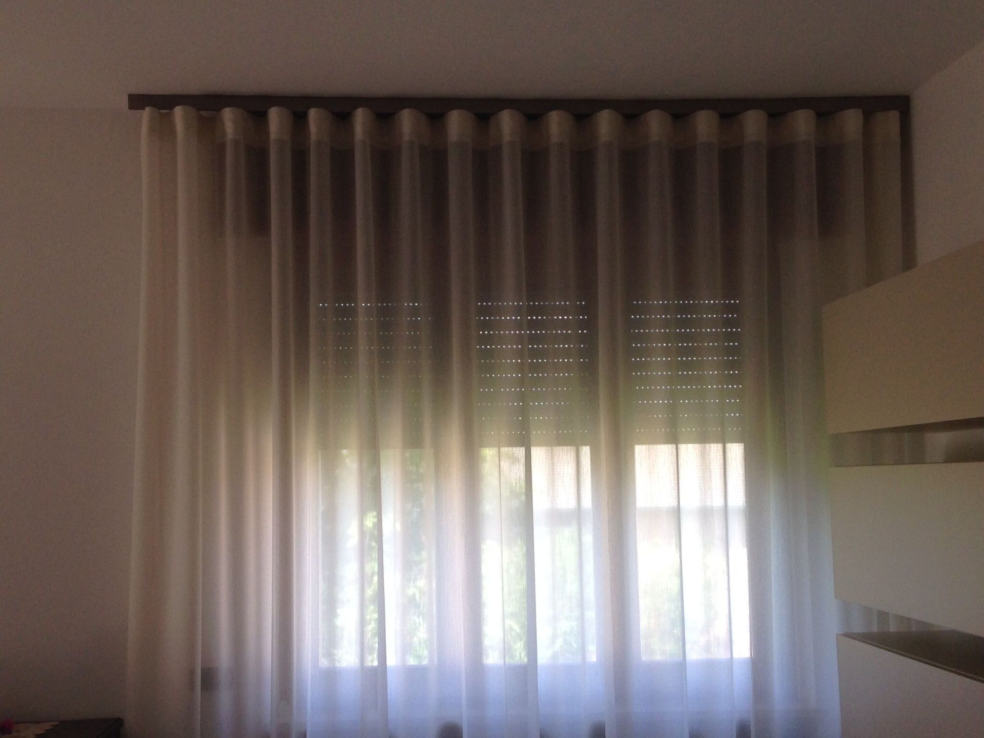 Una finestra coperta da tende leggere e trasparenti color avorio sporco, con pieghe ondulate, appese a un binario superiore.