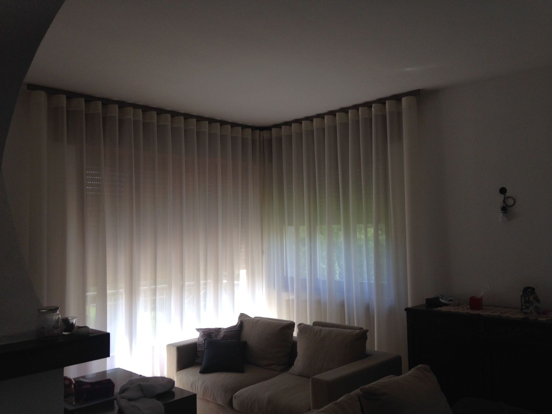 Un angolo della stanza con tende trasparenti di colore chiaro, che arrivano fino al soffitto e coprono due finestre, posizionate dietro un divano.