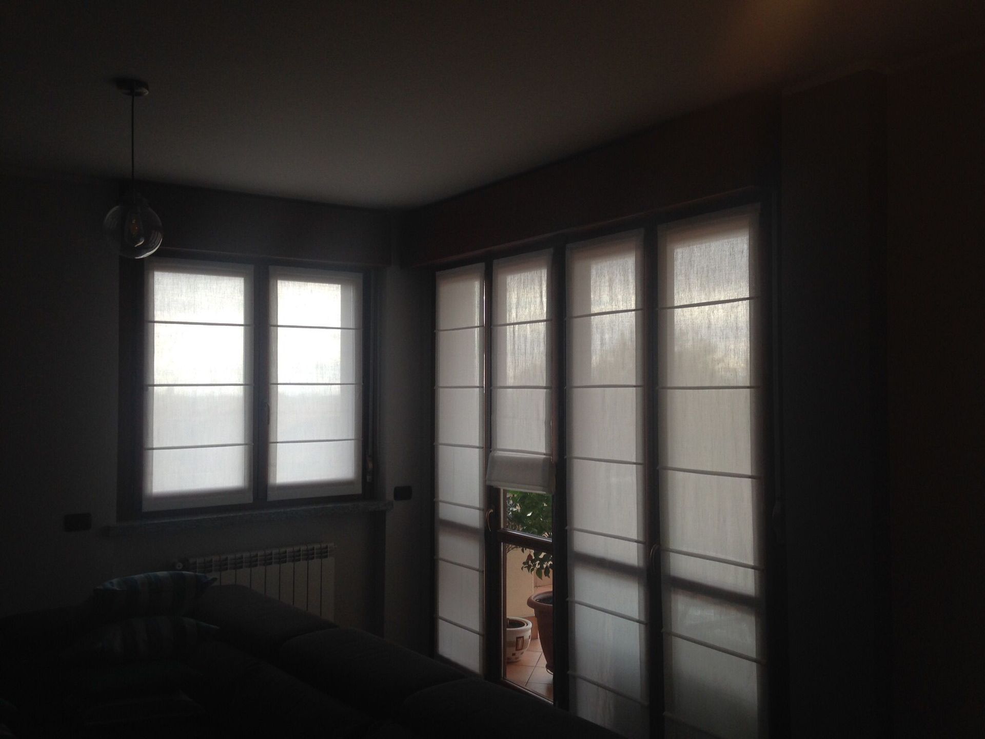 Una stanza scarsamente illuminata con due finestre coperte da tende a pannelli orizzontali, una delle quali si affaccia su un balcone con una piccola pianta in vaso.