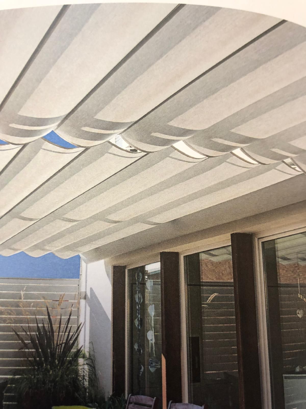 Una tenda parasole retrattile in tessuto bianco copre un patio esterno a una casa con porte a vetri e travi di sostegno in legno.