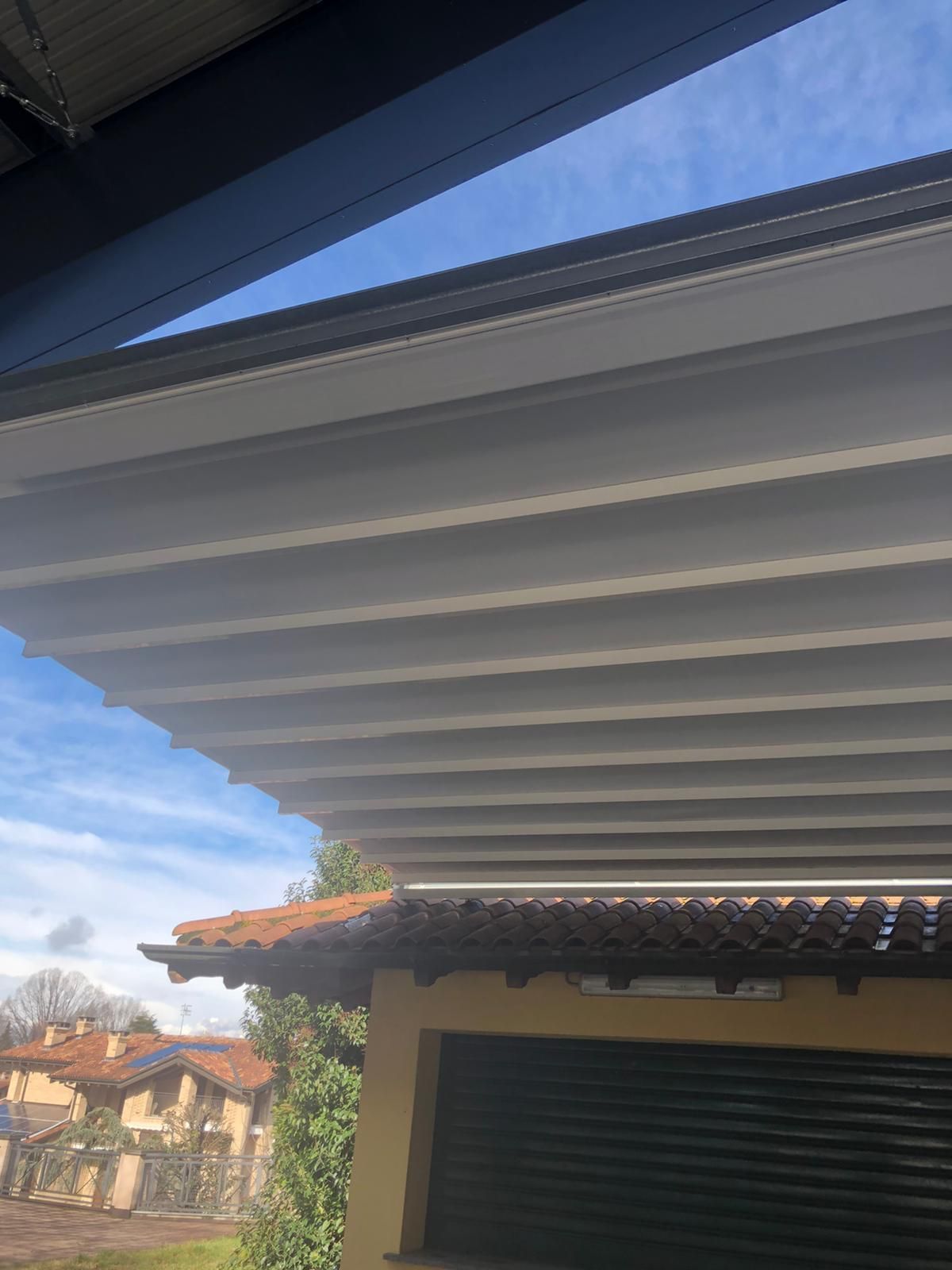 Sotto un cielo azzurro, una tettoia bianca a lamelle si estende sopra un edificio giallo con un tetto di tegole scure e una porta da garage.