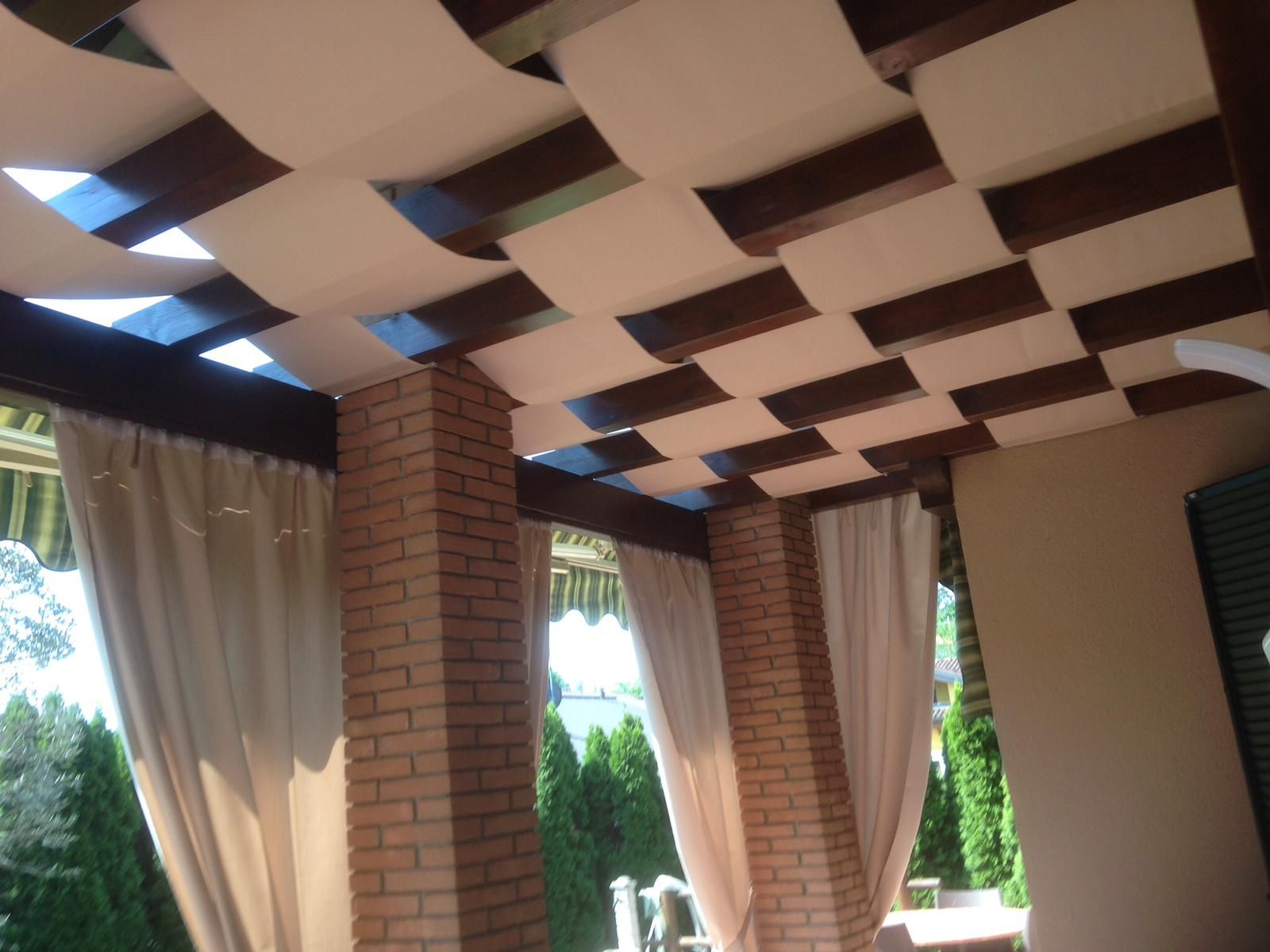 Un patio con un soffitto a pergola in legno caratterizzato da pannelli drappeggiati in tessuto color crema, pilastri in mattoni e tende beige sospese.