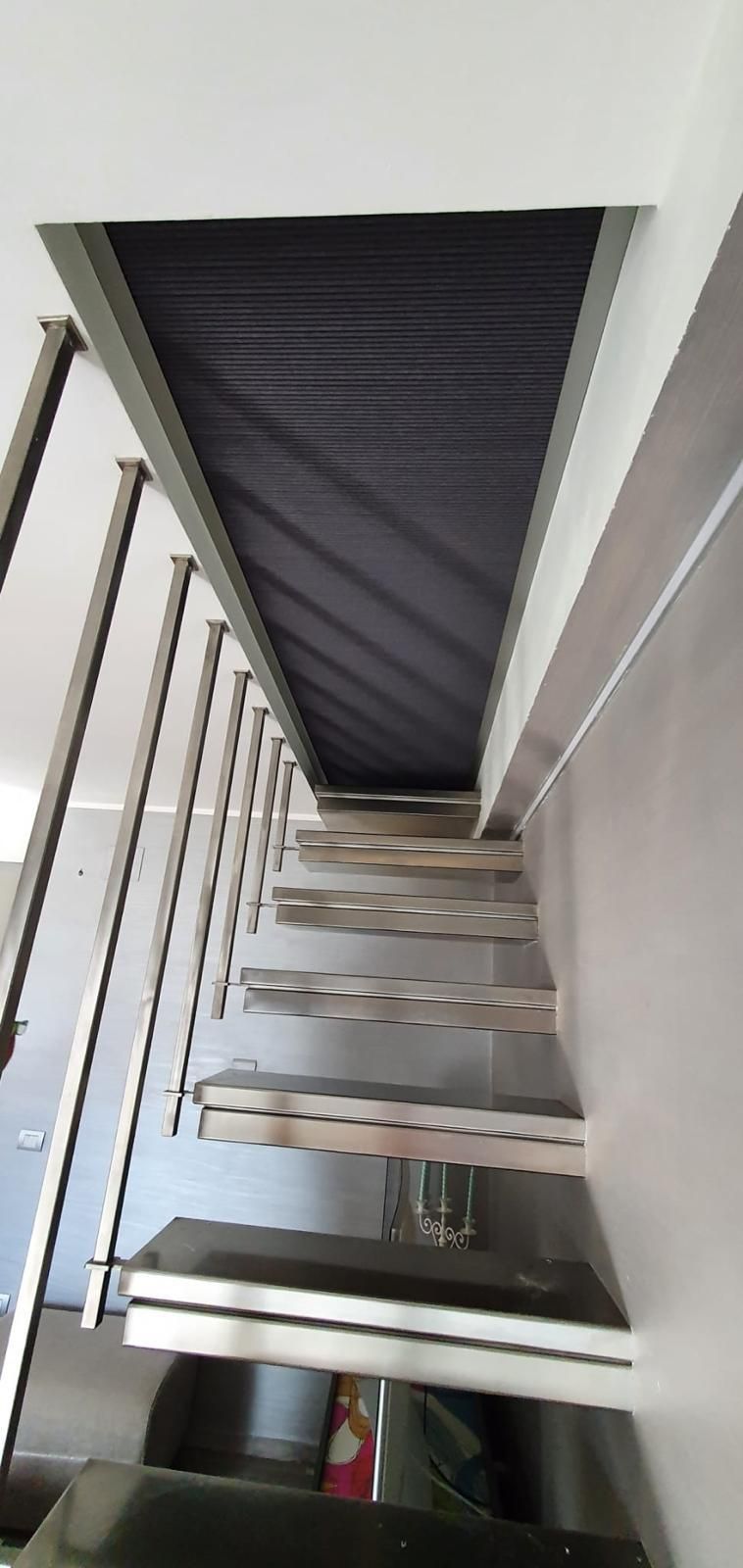 Una scala moderna a vista con ringhiera in metallo che sale verso un'apertura rettangolare scura sul soffitto.