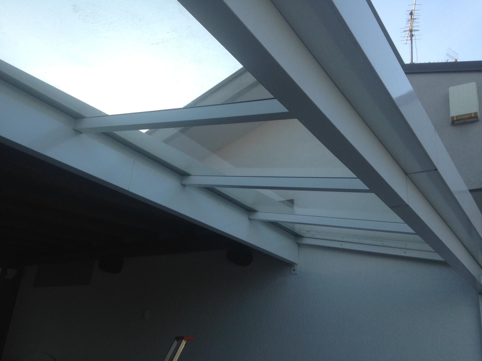 Una struttura di copertura per patio bianca e inclinata, con pannelli traslucidi e travi fissate al lato di un edificio.